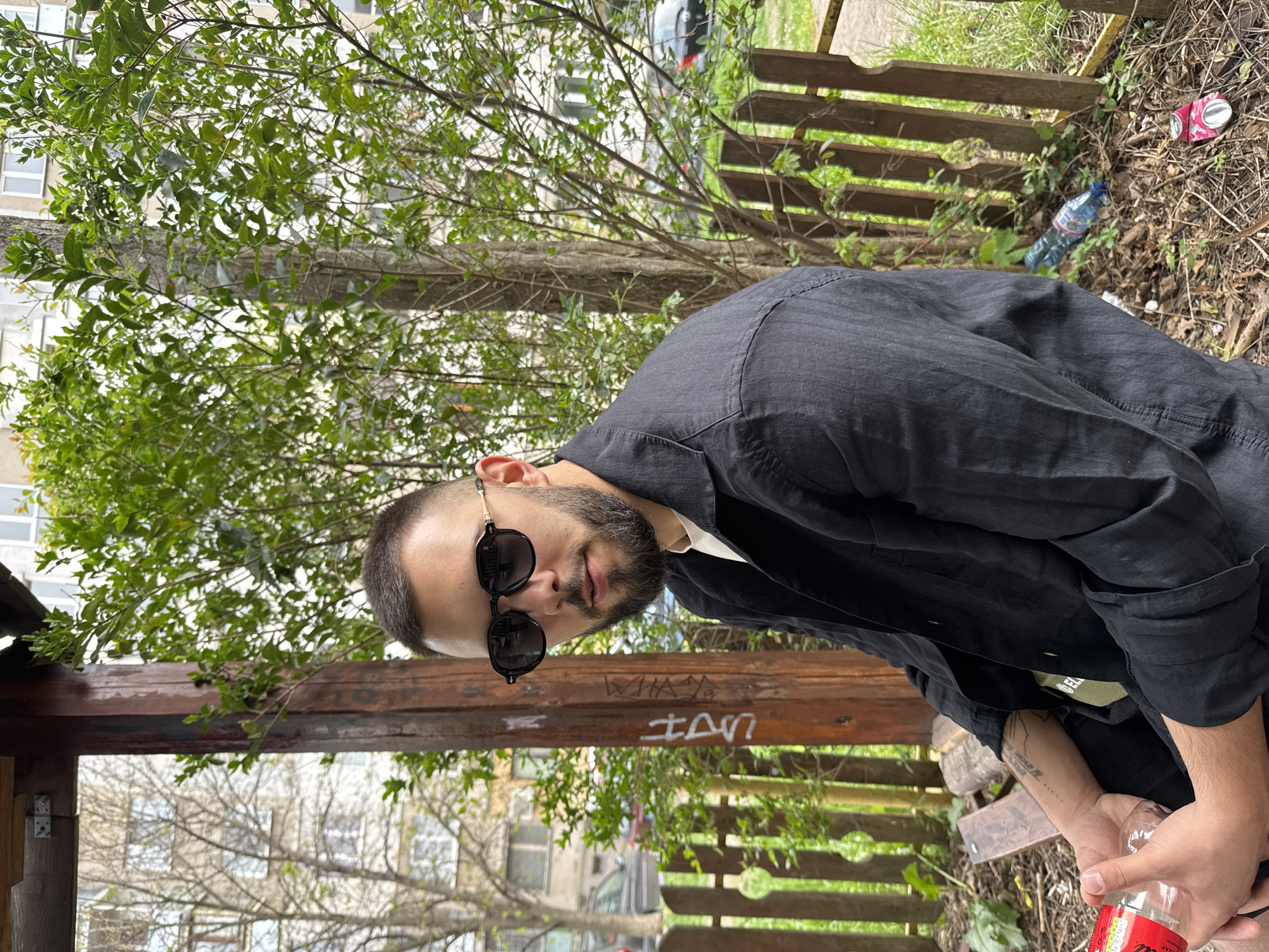 A young man with a beard and short hair, wearing black sunglasses, a black long-sleeved shirt, and a white t-shirt, is looking directly at the camera. He is in an outdoor setting, sitting or crouching beside a sturdy wooden structure, possibly a gazebo or a large bench. He holds a clear plastic bottle with a red label, suggestive of Coca-Cola, in his right hand.
The scene appears to be a somewhat unkempt outdoor area, likely in Sofia, Bulgaria. The ground beneath and around the wooden structure is covered with dirt, dry leaves, and some litter, including a blue plastic bottle and a crushed pink/red aluminum can. Behind the man, green leafy trees and bushes fill the mid-ground, with tall, light-colored residential buildings visible in the background, under what appears to be an overcast sky. A dark car is partially visible among the trees.
Graffiti is etched onto the horizontal wooden beam of the structure, featuring the words "WHAT'S UP" (partially visible) and additional unidentifiable markings below. The lighting indicates it is daytime, but without direct sunlight. The man's expression is neutral, partially hidden by his sunglasses. The overall impression is an informal, urban outdoor moment.