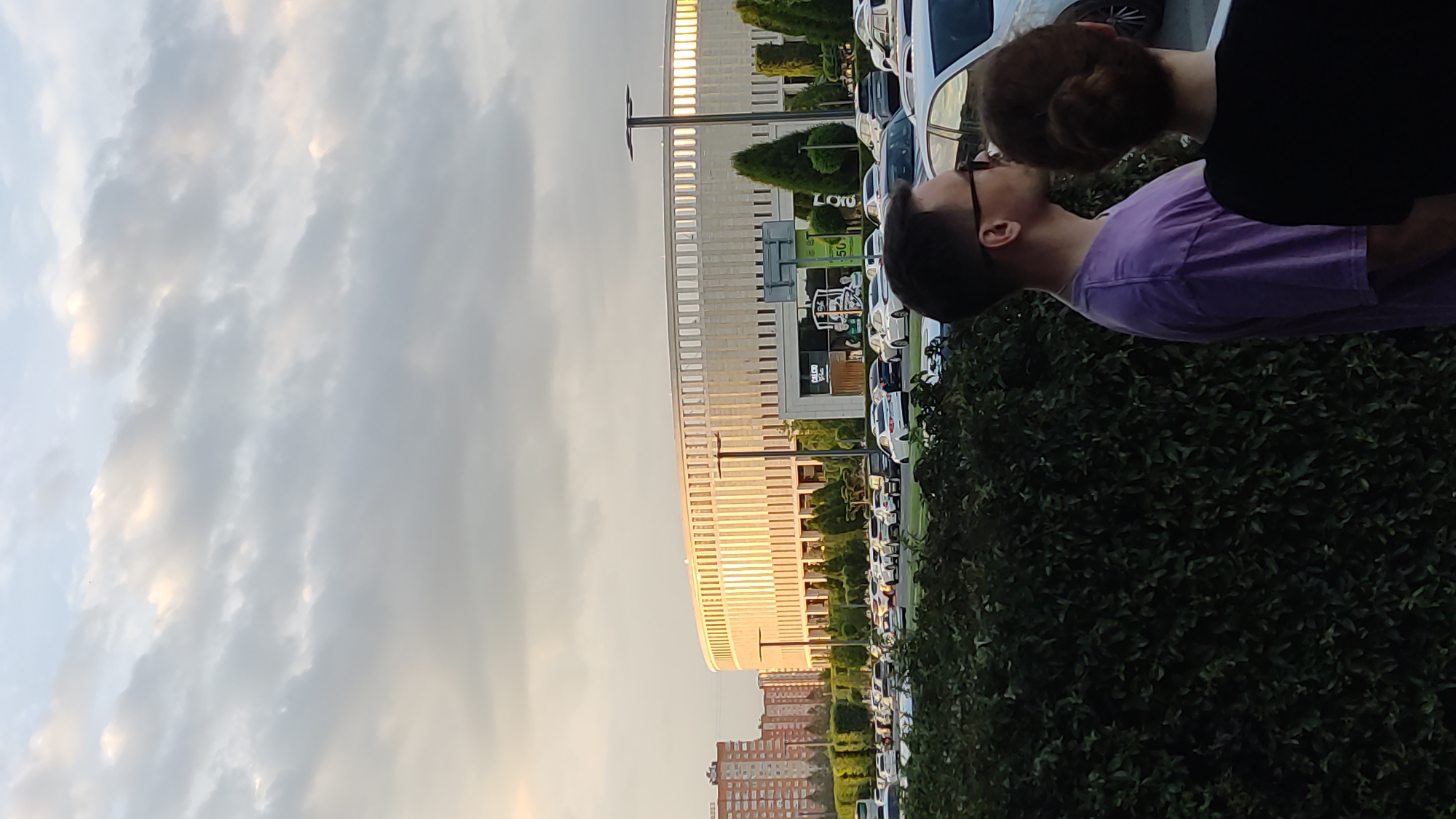 This is an outdoor, eye-level, vertical, medium shot of a building and a parking lot in Krasnodar, Russia. The building, likely a stadium or arena, is circular with a ribbed facade and is illuminated by the setting sun. In the foreground, two people are visible from the shoulders up, facing away from the camera. The person on the left wears a purple t-shirt and sunglasses. The person on the right has their hair in a bun. A dense green hedge is in the immediate foreground. The parking lot is filled with numerous cars, and trees and smaller buildings are visible in the background. The sky is cloudy with patches of sunlight.