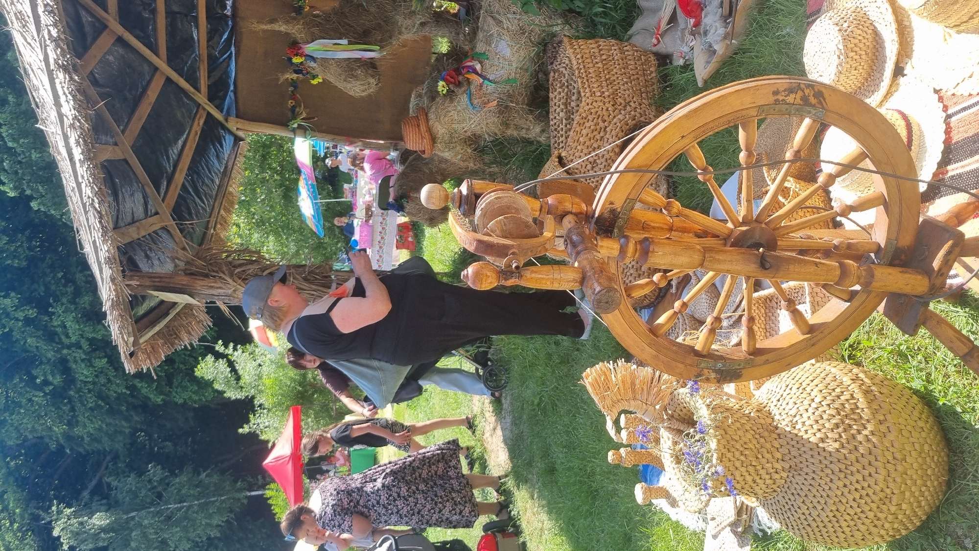 This image captures an outdoor market scene in Melnyky, Ukraine, under bright sunlight. In the foreground, a traditional wooden spinning wheel occupies the lower right and center. To its left, a large woven straw jug-like vase containing dried purple flowers sits on the grass, surrounded by smaller straw craft items. The ground is covered in green grass.
In the midground, a rustic market stall with a thatched roof, partially covered by a black tarp and supported by wooden poles wrapped in natural fibers, serves as a central display. Inside and around this stall, bundles of straw are arranged, some adorned with colorful ribbons and small artificial flowers. One straw bundle in the lower right features a miniature straw hat. A large woven basket rests near these straw displays.
Several individuals are present. A woman wearing a black t-shirt with an orange panel, dark pants, and a grey baseball cap stands with her back partially to the camera, appearing to examine something in her hands near the stall. Behind her, another person in light pants carries a large light blue bag. To the left, a woman in a patterned dress is visible, and further left, a child's head is partially seen. In the background, under a red tent and various colorful striped umbrellas, more people are observable at other market stations. The background is dominated by dense green foliage, suggesting a wooded area or forest.