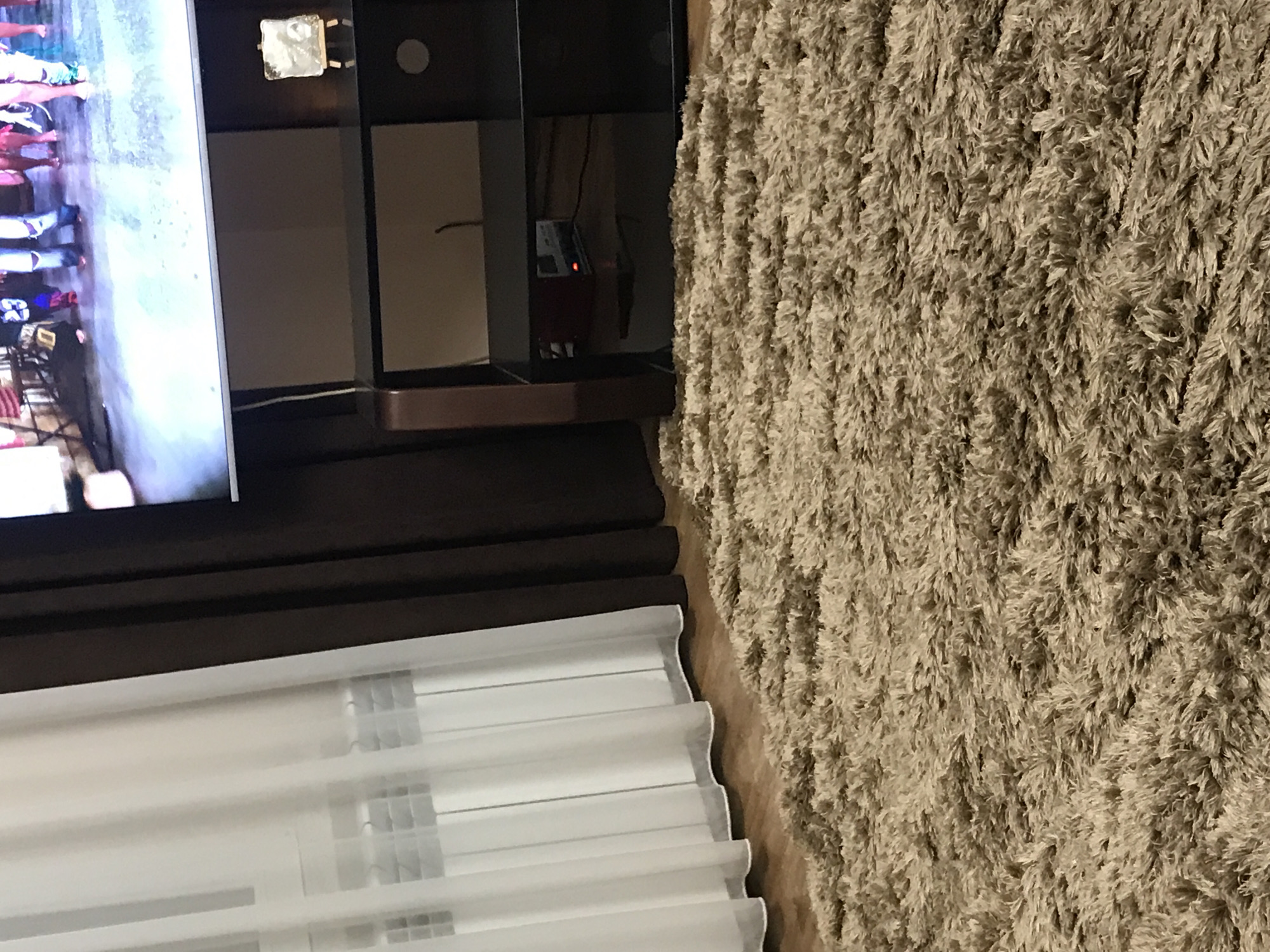 The image shows an interior setting. On the left, there's a television screen displaying figures and objects, partially obscured by a black TV stand. Below the TV stand are white, pleated blinds. Adjacent to the TV stand is a tan shag rug. The scene appears to be indoors. There is no information about Yerevan, Armenia, or any people.
