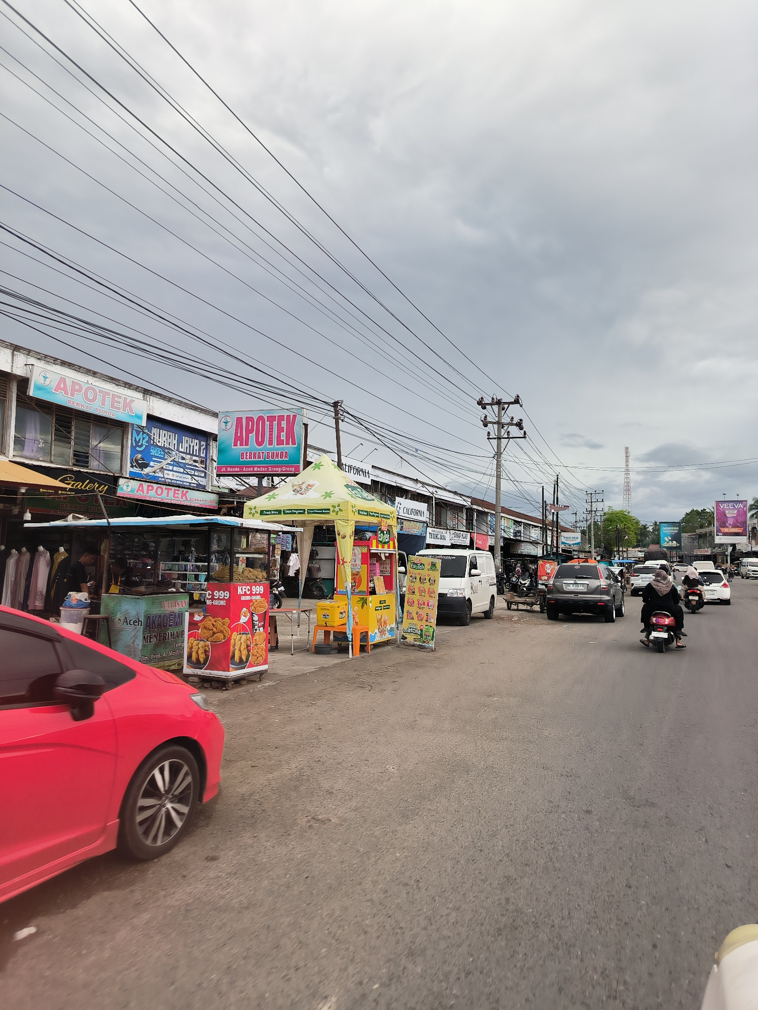 The image presents a busy street scene in Grong-Grong, Indonesia, under an overcast sky. In the foreground on the left, a red car is partially visible, either parked or in motion. Adjacent to the road, a row of commercial buildings lines the street, featuring various shops and businesses.
Prominently displayed are multiple signs for "APOTEK BERKAT BUNDA," with one sign specifying its location as "Jl. Banda Aceh Medan Grong-Grong." Other visible business names include "AURAH JAYA 2," "Galery," "KALIFORNIA," "TRINA JAYA," and an "Aceh AKADEMI" sign indicating student enrollment.
On the sidewalk, several street vendors operate under vibrant yellow and green tents. One stall features "KFC 999" branding, displaying fried chicken and other items. Another stall offers drinks and snacks, including "matcha," with text like "Fresh Brew" and "Kopi Aceh." A person can be seen working at one of the stalls.
The paved road in the mid-ground shows moderate traffic, including a white van, a grey SUV with license plate "BL 1199 PR," a white sedan, and several motorcycles and scooters. One rider on a scooter is visible wearing a headscarf.
The sky is a uniform grey, indicating cloudy weather. A dense network of black utility lines and cables crisscrosses overhead, supported by tall utility poles. In the distance, a radio or telecommunications tower stands against the horizon, and billboards, including one for "VEEVA," are visible further down the street. The scene depicts a lively commercial area with ongoing daily activities.