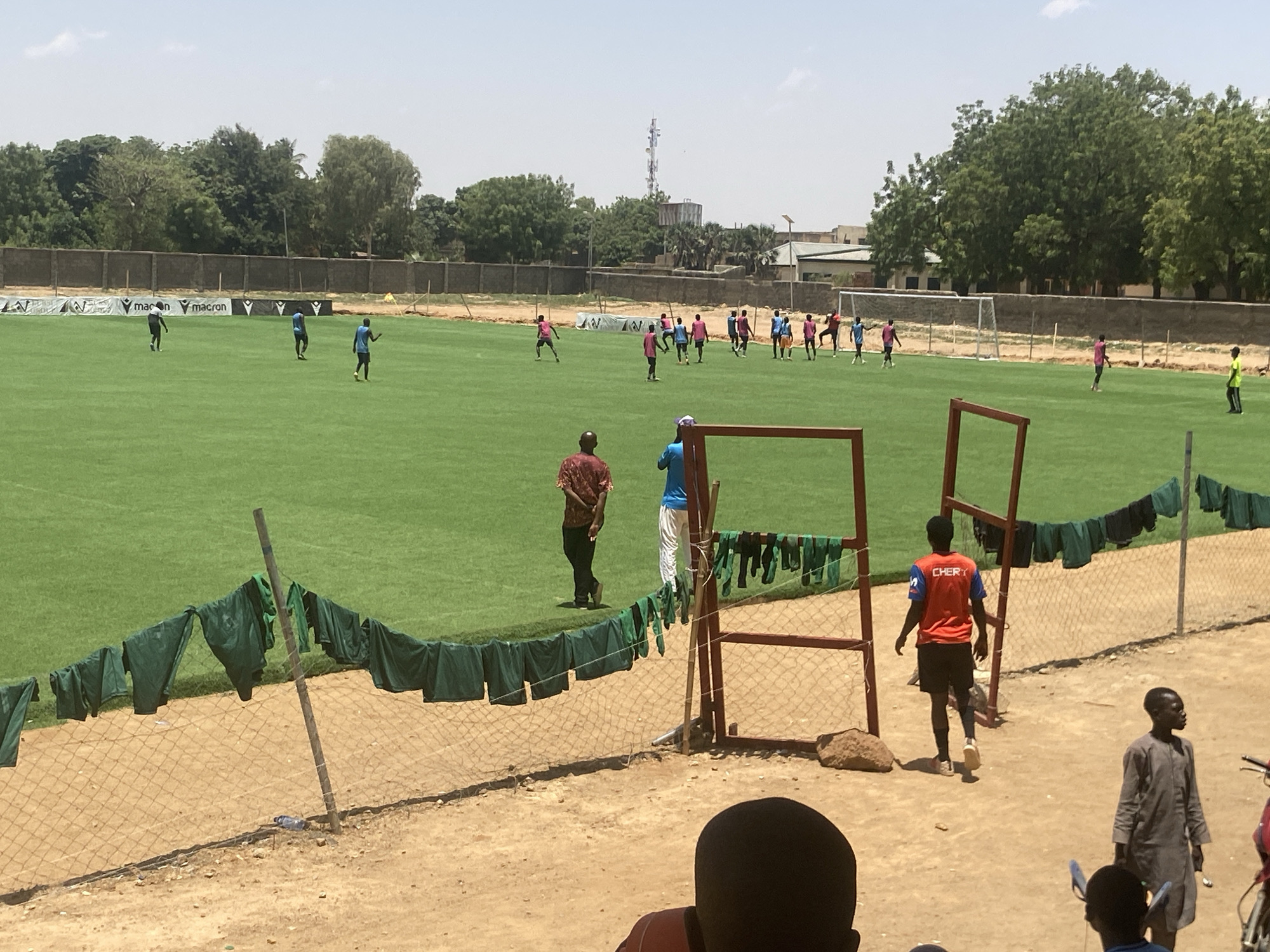 The image shows a soccer field in Tudun Wada, Nigeria, with players in various colored jerseys scattered across the green field. A group of players wearing pink and blue jerseys are gathered near the goal. Two men stand near a rusty red metal frame, one in a brown patterned shirt and the other in a blue shirt and white pants. Green shirts are draped along the fence bordering the field. A player in a red shirt with the word "CHERY" on the back walks away from the field. A man in a gray shirt walks alongside the fence. The ground surrounding the field is dirt. Buildings and trees can be seen in the background. A communications tower is visible in the distance.