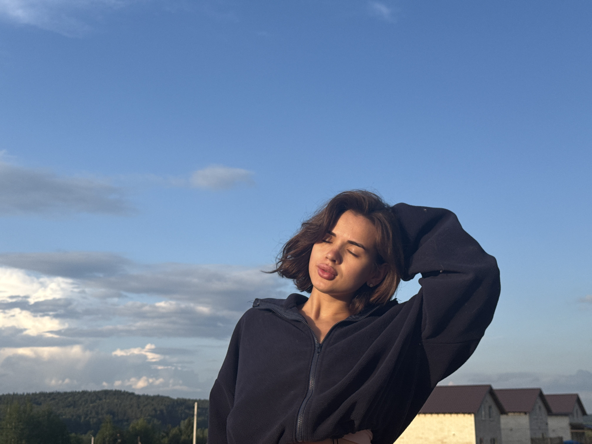 A young woman with short brown hair, wearing a navy blue zip-up hoodie, stands outdoors in what appears to be the city of Lobanovo, Russia. Her eyes are closed and she purses her lips slightly, and one hand is raised near her head. Behind her, the scene features a clear blue sky with scattered white clouds. The horizon reveals a treeline, suggesting a wooded area. In the lower right corner, there are three small, light-colored buildings with brown roofs, possibly residential homes. The photo appears to be taken during the daytime, likely during a bright afternoon.