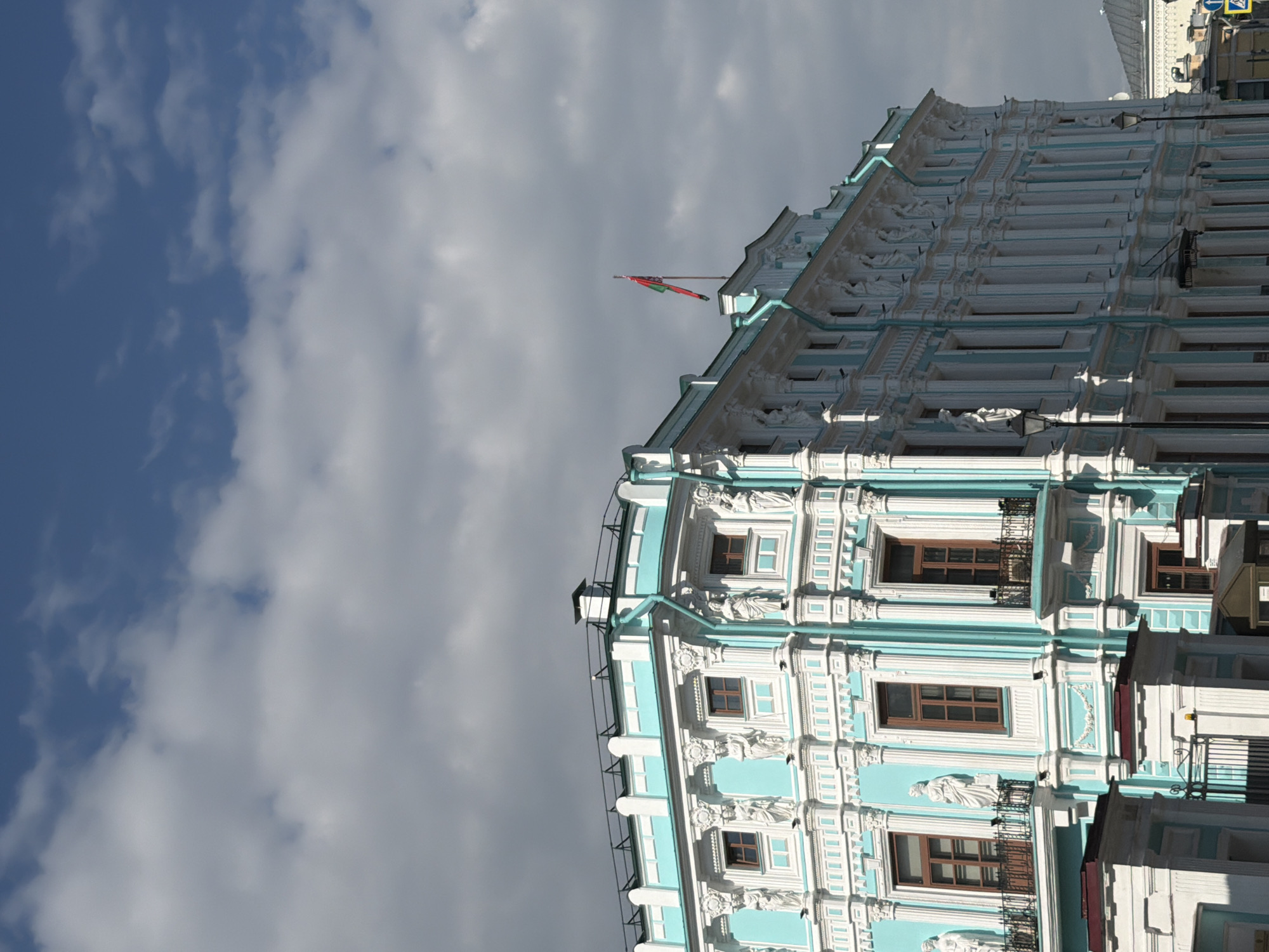 The image shows a building in Moscow, Russia, specifically the Pashkov House, a neoclassical mansion. The building has a pale blue facade and intricate white ornamentation, including statues and columns. Windows are visible with dark frames. A red and green flag flies atop the building, possibly the Belarusian flag. The sky is visible in the background, with blue patches interspersed with white clouds. The overall view is from a low angle, emphasizing the height and architectural details of the building. The image is rotated counter-clockwise.