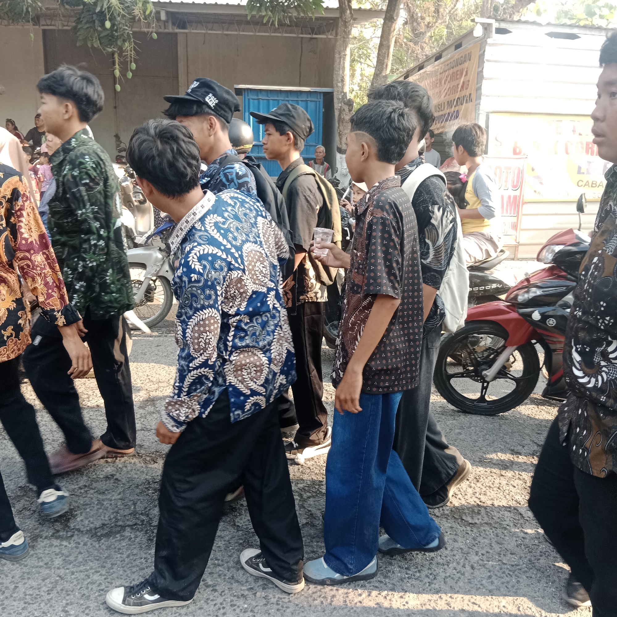The image shows a group of young men walking in a procession on a paved street in Tegalrejo, Indonesia. The men are predominantly wearing batik shirts and dark pants. Several individuals are wearing black caps. In the background, there are buildings, trees, and motorcycles. One man in the foreground is holding a clear plastic cup. The environment appears to be outdoors, with sunlight casting shadows on the ground. The overall scene suggests a casual outdoor event or gathering.