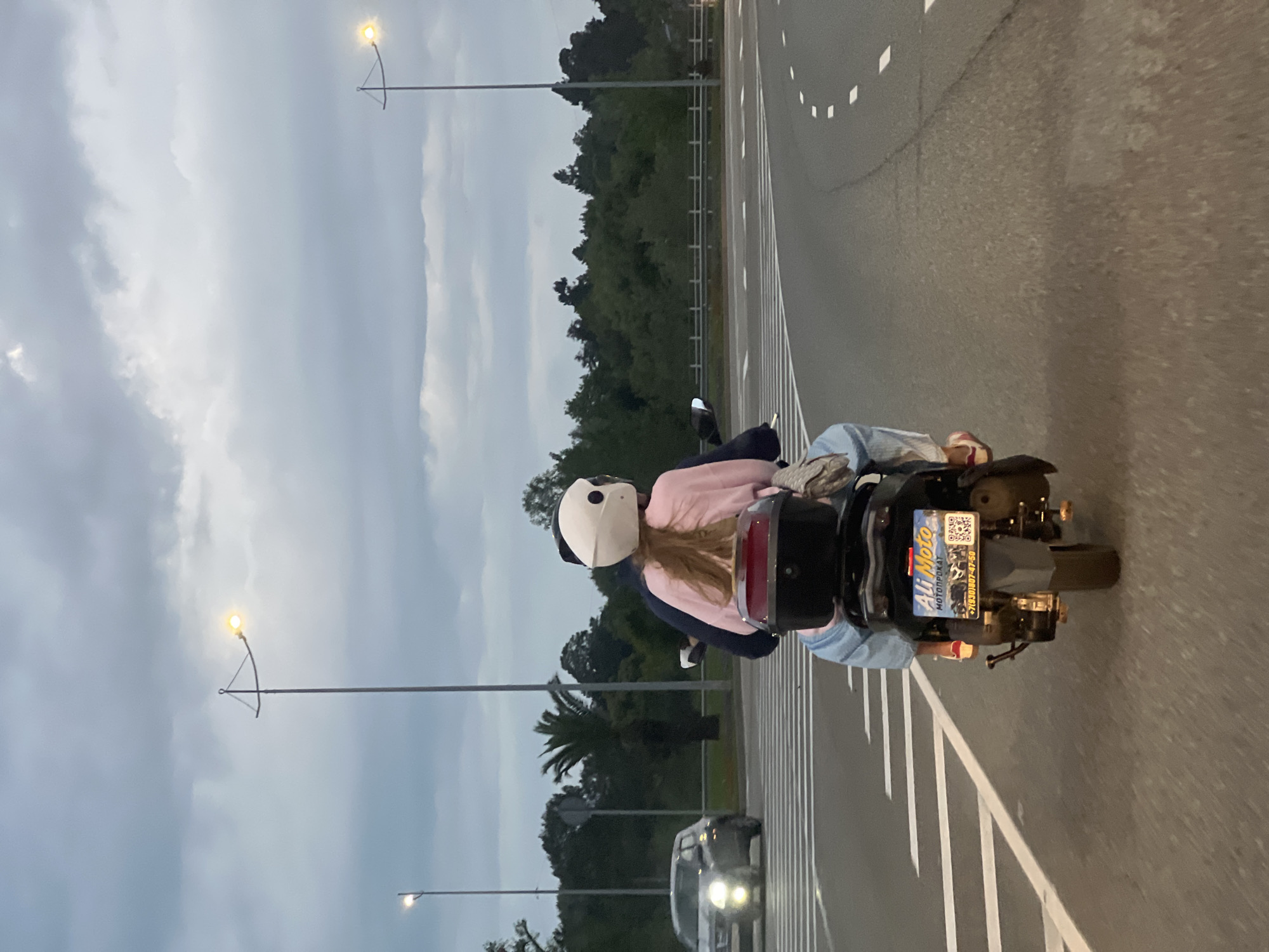 A person wearing a white helmet and a pink jacket is riding on a moped on an asphalt road. The moped has a license plate with Cyrillic script, including "Ali Moto". Another person is seated behind. The road has white lane markings. Street lights line the side of the road. Trees are visible in the background. A vehicle is driving behind the moped. This photograph appears to be taken in Sochi, Russia.