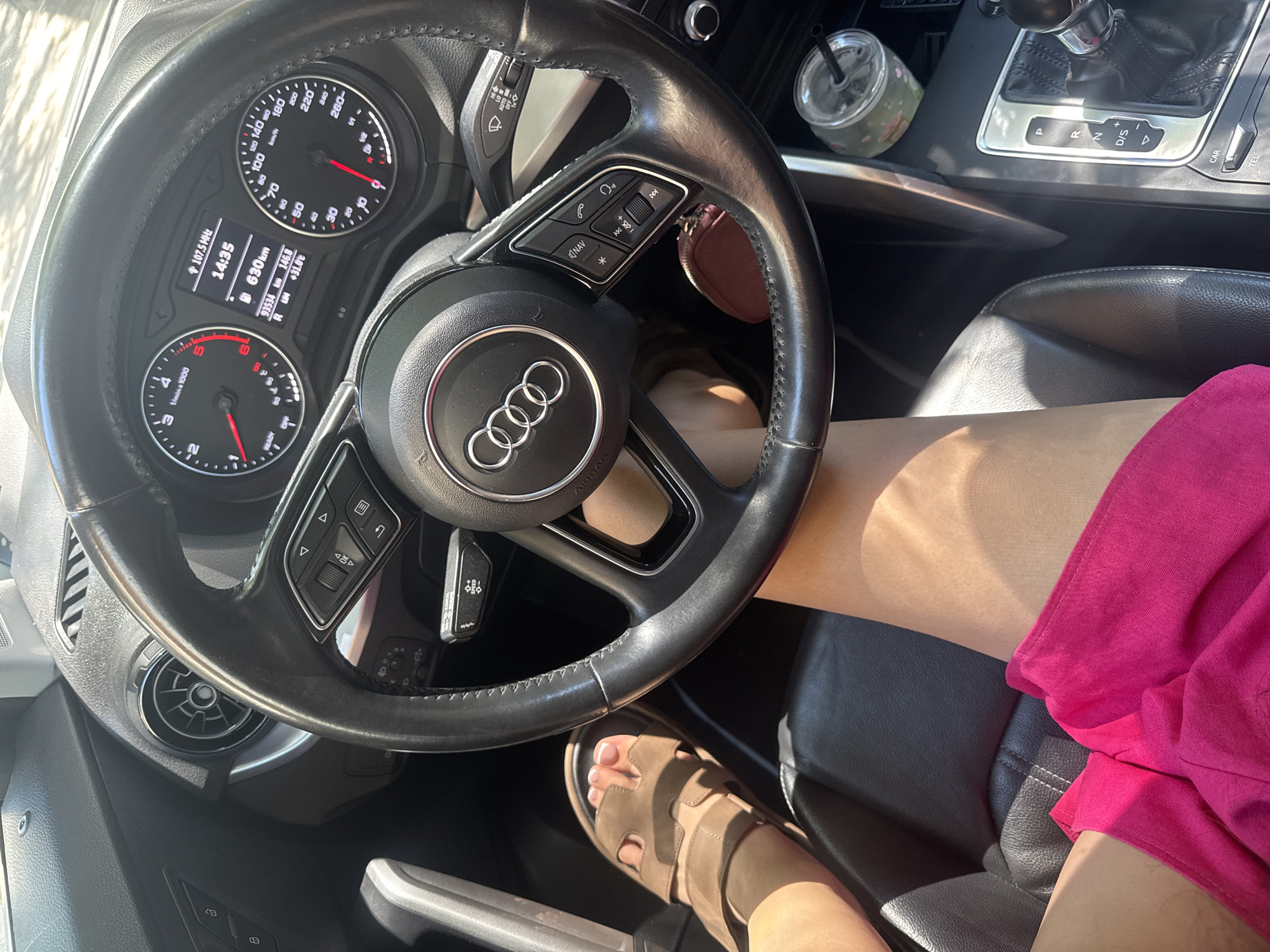The image shows a close-up view of the interior of a car, specifically the driver's side. The steering wheel, dashboard with an instrument cluster displaying speed, RPM, fuel level, and trip information, and the gear shifter are visible. A person's legs are in the frame, with one foot wearing a tan sandal, suggesting they are the driver or a passenger. A bright pink garment is visible on the person's lap. The overall scene suggests the vehicle is parked or in motion, likely on a sunny day given the lighting and shadows. There is no visual information to determine the specific location within the city of Plovdiv, Bulgaria, or any urban features.