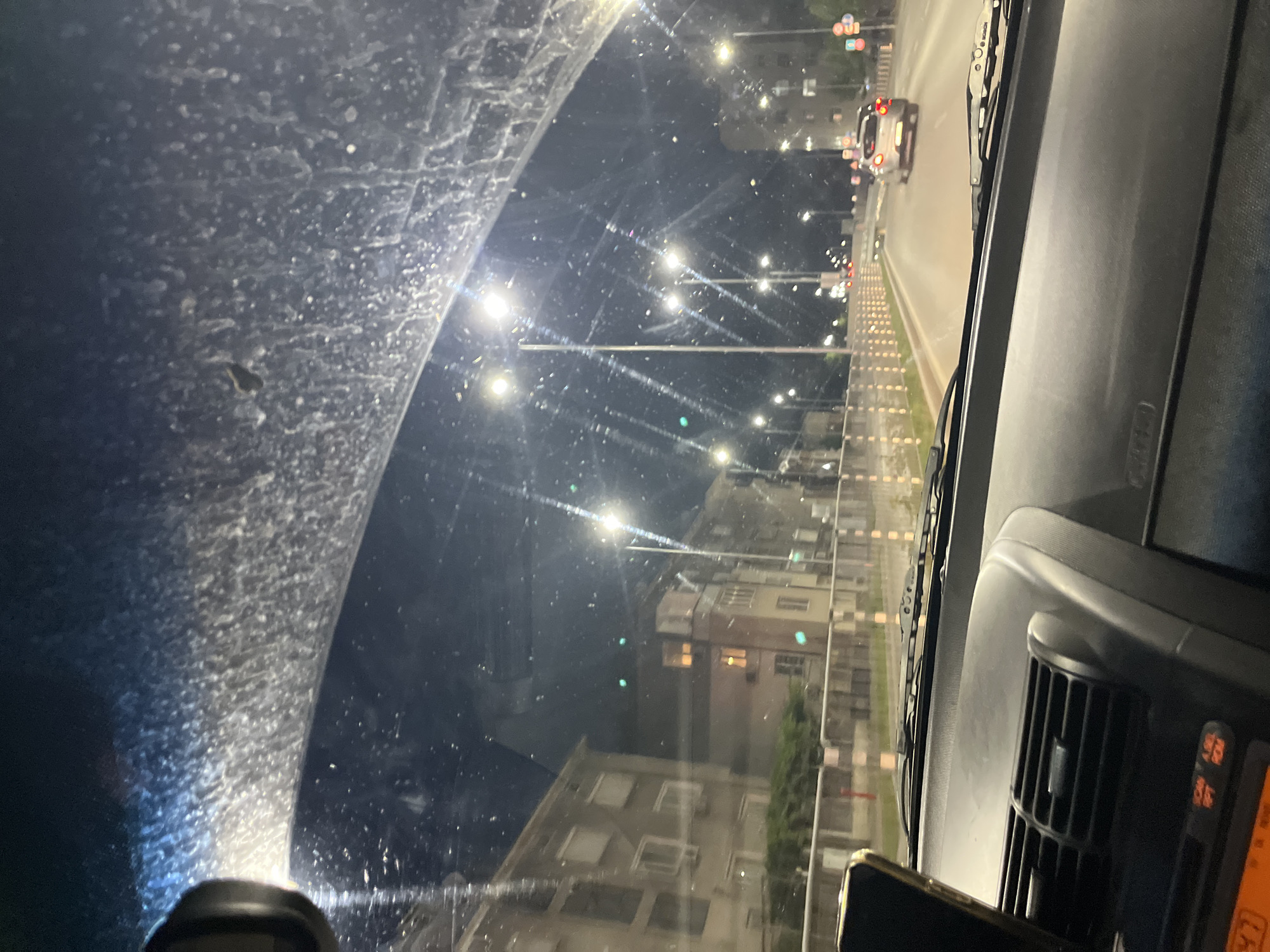 This is a first-person perspective shot from inside a vehicle at night, likely Plovdiv, Bulgaria, as indicated by the location context. The windshield is dirty and covered in water spots, creating a diffused and somewhat obscured view of the outside.
The scene outside is a street at night, illuminated by streetlights and the headlights of passing cars. The road is lined with what appear to be reflective markers or small lights. Buildings with illuminated windows are visible along the street, suggesting a moderately urban or suburban environment. A white car with its brake lights on is visible in the distance, driving away from the viewer. The overall impression is of driving through a city at night.
The time of day is clearly night, and the presence of water spots on the windshield suggests it might be raining or has recently rained. The lighting is typical of a city at night, with bright streetlights creating glare and reflections.
Notable details include the dirty windshield, the line of lights along the road, and the illuminated buildings. The interior of the car is partially visible, with a dashboard and what appears to be a phone mounted near the center console. There is some faint orange text visible on a car radio display or similar device. No people are explicitly visible in the frame, but the viewpoint implies the presence of a driver. The mood conveyed is one of travel and observation during a nighttime drive, with the dirty windshield adding a slightly gritty or atmospheric element.