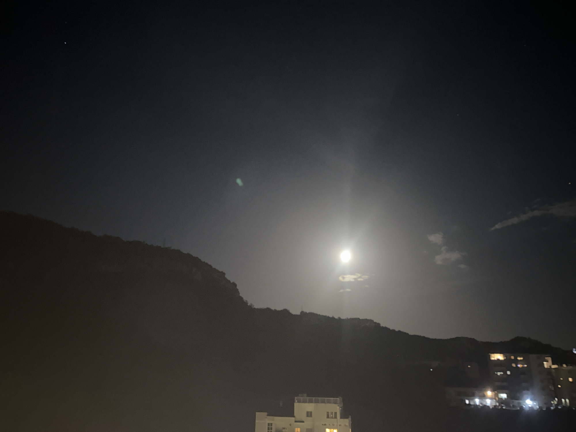 A night scene in Rafailovići, Montenegro, features a bright full moon illuminating the sky above a silhouetted mountain range. The moon, positioned slightly right of center, casts a luminous glow and creates lens flare. A few wispy clouds are visible near the moon. Below the mountain, a town with illuminated buildings is visible. One building, appearing to be a multi-story apartment complex or hotel, is in the foreground, while other structures with lights are spread out in the background along the coast. The overall atmosphere is dark, with the moon and building lights serving as the primary light sources.