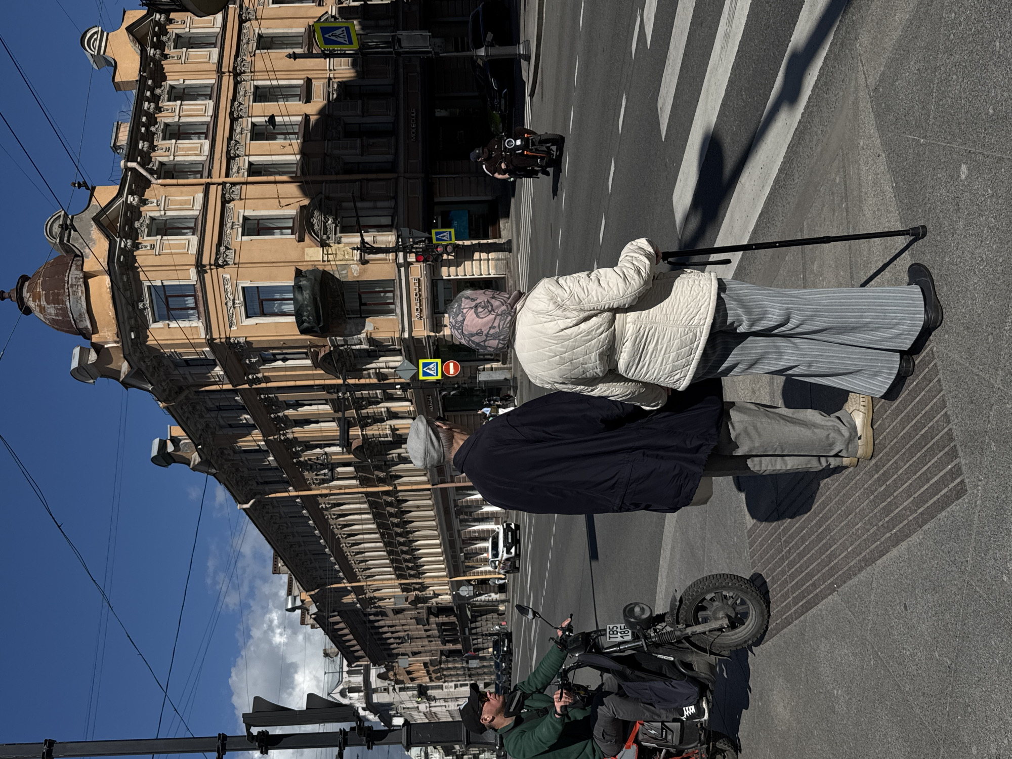 This is a street-level photograph taken outdoors on a sunny day, with the camera positioned vertically. The scene depicts a city street with classic European architecture. The buildings, painted in warm beige and ochre tones, feature multiple stories with ornate detailing and numerous windows. A clear blue sky with a few wispy clouds is visible above.
The foreground is dominated by a pedestrian crossing with white stripes on asphalt. Two individuals are in the frame, standing on the sidewalk near the crossing. One person, viewed from the back, wears a quilted white jacket and striped grey pants, and holds a dark walking stick. Their head is covered with a patterned scarf or headwrap. Next to them, also viewed from behind, is another person wearing a dark jacket and grey pants.
Further down the street, a motorcycle is parked, and a person wearing a green jacket and a baseball cap is seen near it, looking upwards. In the background, a few vehicles, including what appears to be a white car and a motorcycle with a rider, are visible on the street. Traffic signals and pedestrian crossing signs are also present.
Shadows cast by the buildings and the people suggest it is daytime, likely late morning or early afternoon given the angle of the sun. The overall atmosphere appears calm and urban. There is some visible text on signs, such as a yellow pedestrian crossing sign with a blue square and a white symbol, and on the license plate of the motorcycle which reads "TBS 18F". Another sign on a building appears to have the word "MOLECULE" written on it vertically.