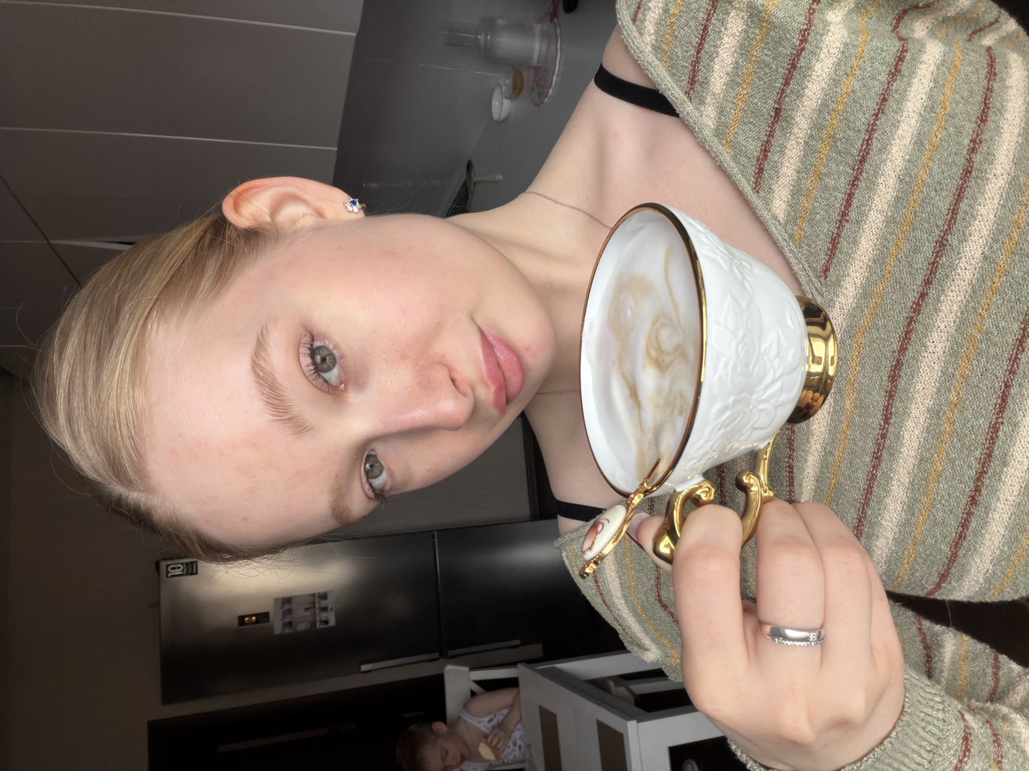 A young woman is depicted in a close-up, slightly tilted portrait, holding a decorative cup filled with a beverage featuring latte art. She has light blonde hair, which is pulled back, and striking green-grey eyes. Her expression is neutral, looking directly at the camera with her lips slightly parted. She is wearing a light green sweater with horizontal stripes in beige, light brown, and orange hues, and a black strap is visible underneath, possibly from a bra or tank top. A delicate silver-toned necklace adorns her neck. On her left ear, she wears a small blue flower-shaped earring, and on her left ring finger, a silver-colored ring with small stones is visible.
The cup she holds is white with a textured design, featuring gold trim around the rim and a prominent gold handle with an ornate, possibly floral, detail. The background suggests an indoor setting, likely a kitchen or dining area, with modern, dark-toned appliances visible, possibly a refrigerator, on which a "10" is discernable. In the lower background, a young child is seated in a light-colored high chair or similar piece of furniture, appearing to look down or be engrossed with an object in their hands. The lighting suggests daytime. There are no specific cues within the image itself to confirm the location as Penza, Russia.