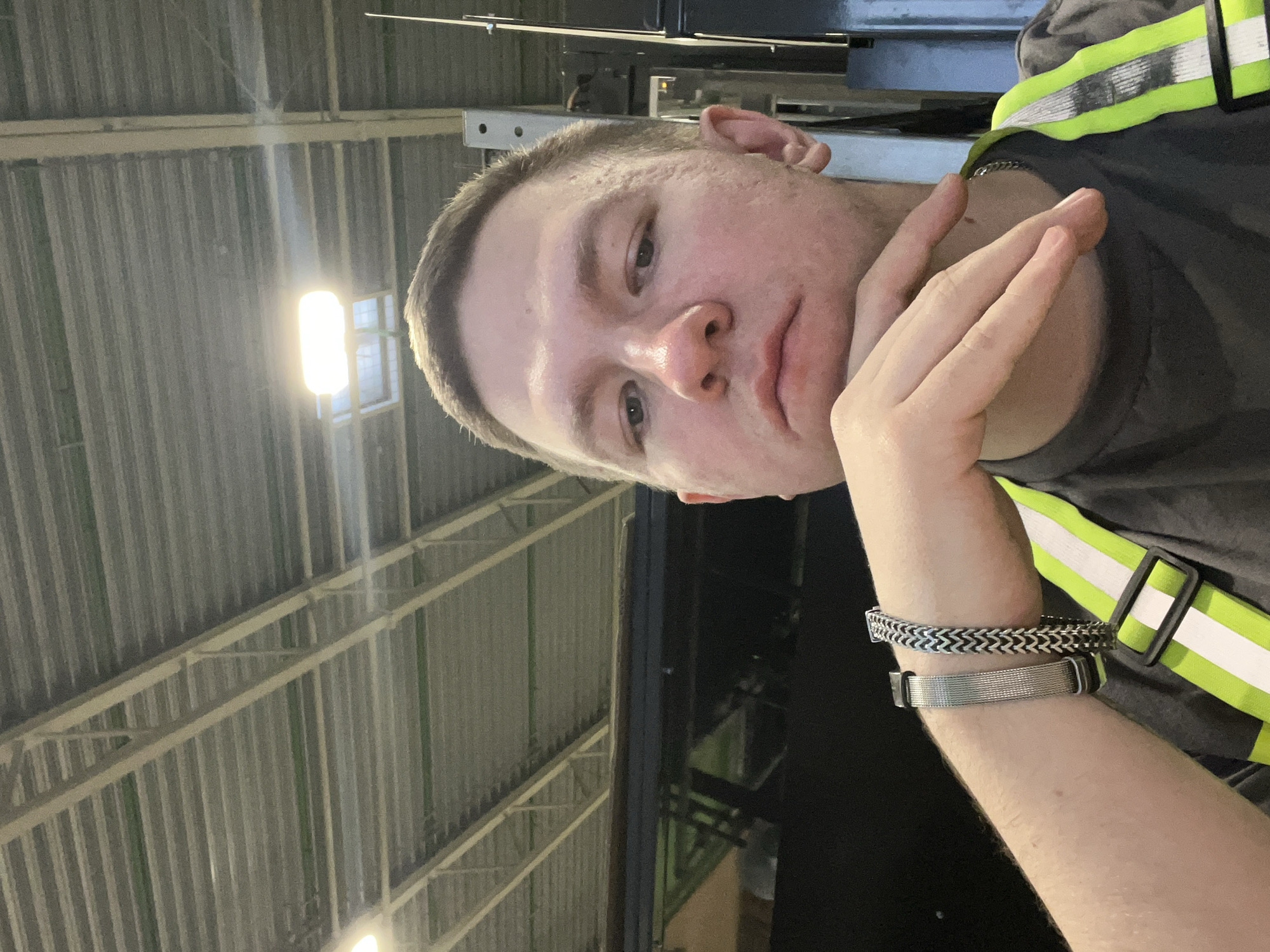 The image is a selfie taken by a light-skinned male with short brown hair. He is wearing a dark grey t-shirt and a neon green and white reflective vest. He has two silver bracelets on his left wrist and is resting his chin on his right hand, with his fingers extended. The background shows the corrugated metal ceiling of an industrial or warehouse-like building, illuminated by fluorescent lights. There is also a dark structure, possibly a vehicle or equipment, visible behind him. The location is identified as Lupanovo, Russia.