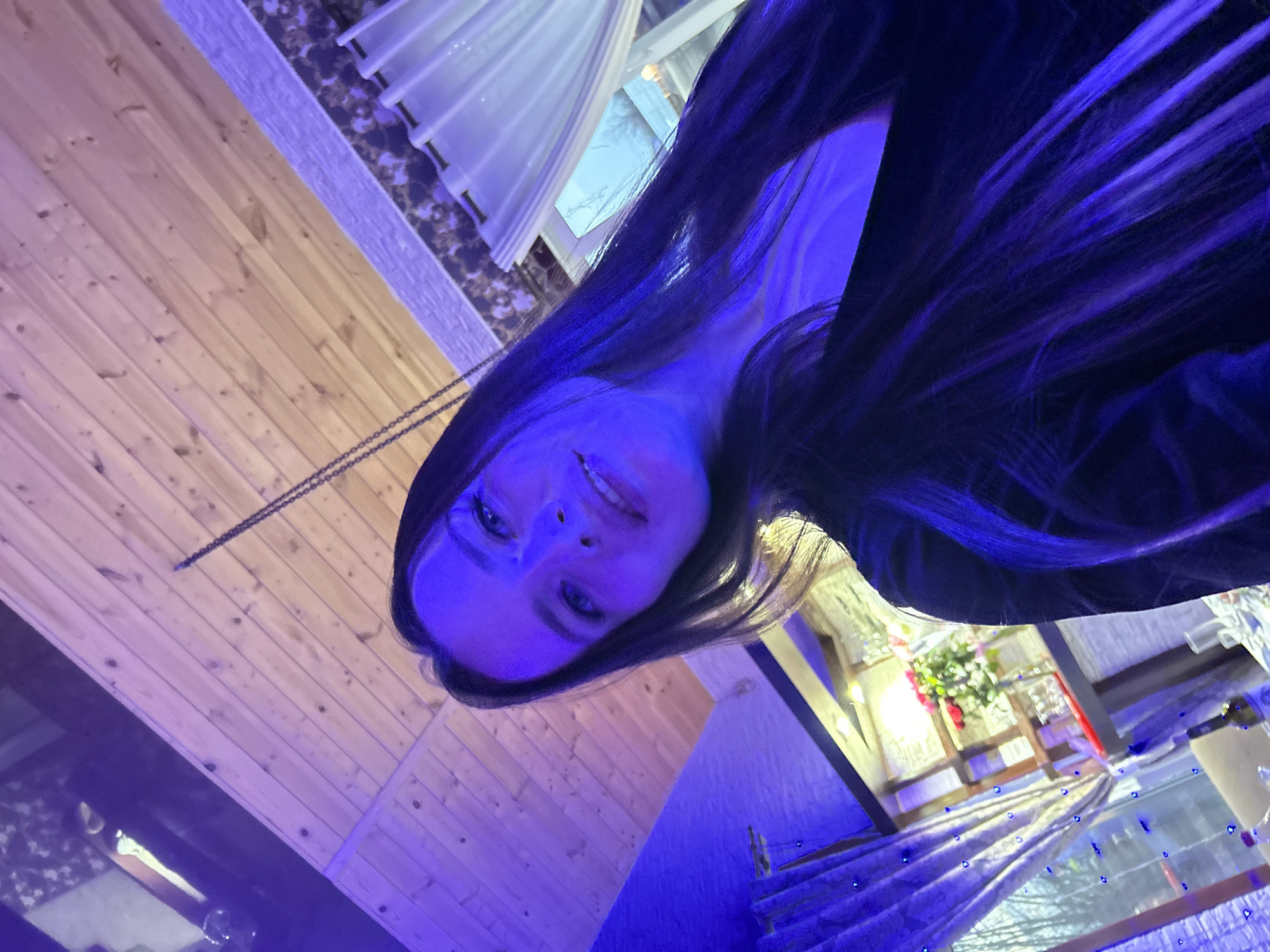 An indoor portrait captures a young woman in a setting bathed in a strong blue-purple light, likely in Chaikovsky, Russia. The woman, with long dark hair tinted blue by the artificial illumination, looks directly at the camera with a faint smile. Her face is clearly visible, with her eyes and facial features highlighted by the cool lighting.
The background suggests an intimate yet stylish environment, possibly a bar, lounge, or restaurant. To the left, a wooden plank wall or ceiling adds a rustic element. Above, a white, translucent curtain or fabric hangs. Further back, a brighter area is visible, possibly a window or a significant light source, accompanied by hints of green foliage and red floral decorations. A thin chain dangles from the wooden structure. Below, a reflective surface, possibly a table, shows numerous small blue light points, echoing the overall blue ambiance. The pervasive blue lighting suggests the photo was taken at night or in a dimly lit space designed for atmosphere. No discernible text is visible in the image.