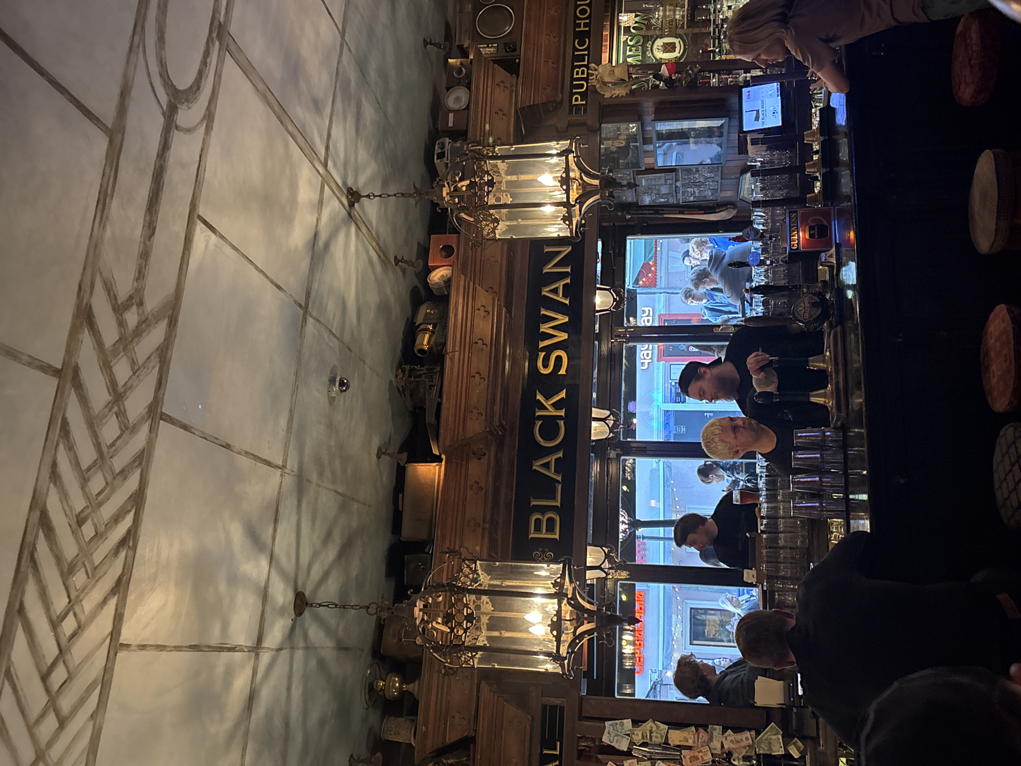 The image shows an interior view of the Black Swan Public House in Moscow, Russia. The bar counter is visible with people standing and sitting around it. There are glass racks above the bar. A sign reads "Black Swan" in gold letters. Above that are some decorations and the sign of the bar says "PUBLIC HOL". Chandeliers are visible hanging from the ceiling. The wall design appears to be an art deco motif. Cash and receipts can be seen on the bar's front.