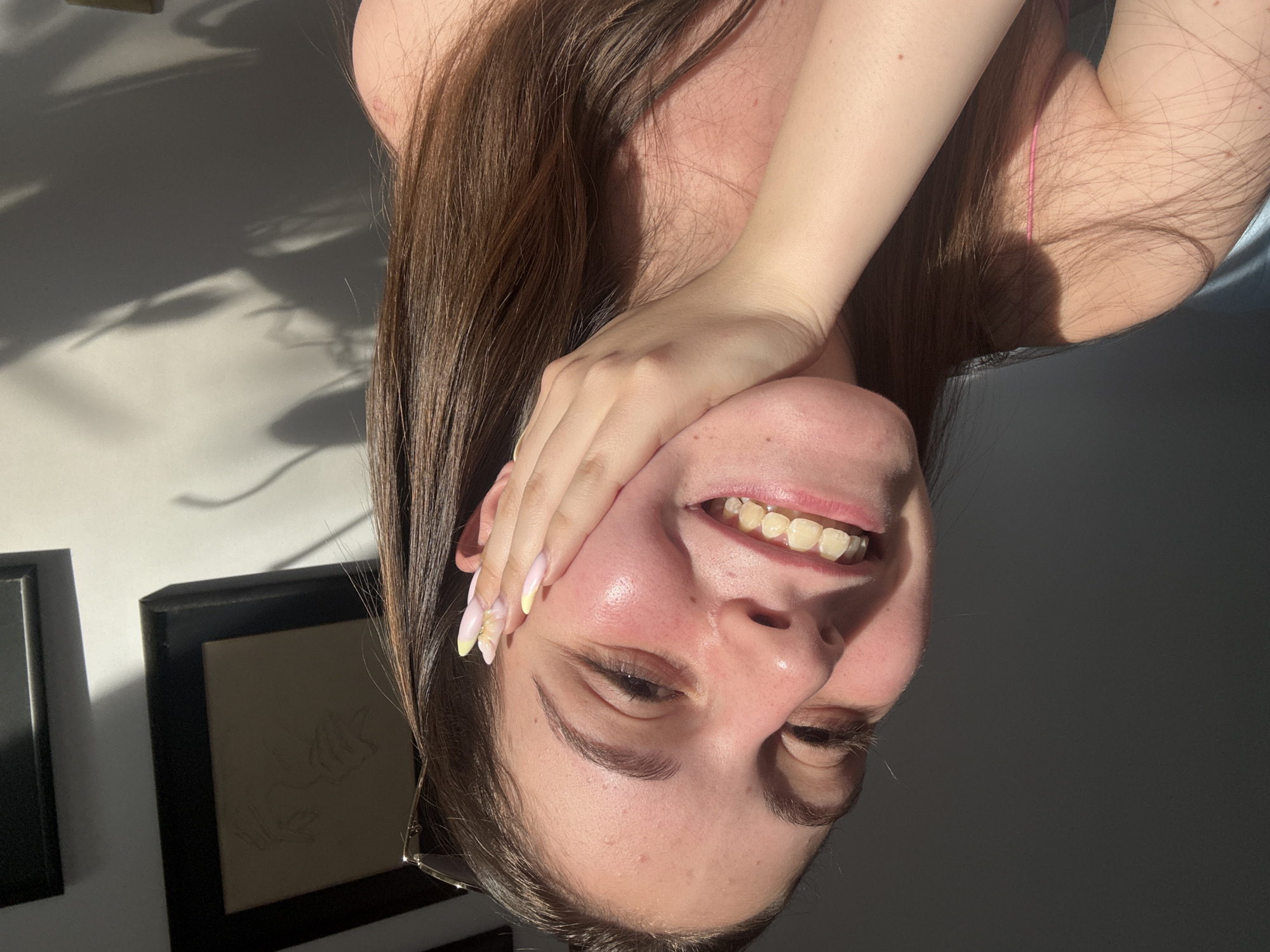 A young person with brown hair and fair skin is captured in a close-up selfie. They are smiling, with their right hand resting on their cheek, and their nails are painted with a light pink and yellow gradient. The subject is wearing a thin pink strap. The background is a plain wall with shadows cast by what appear to be leaves. Framed artwork hangs on the wall. The lighting suggests it is daytime, with sunlight creating highlights on the subject's face and casting strong shadows. The image does not provide any specific information about the location in the city of Burgas, Bulgaria.