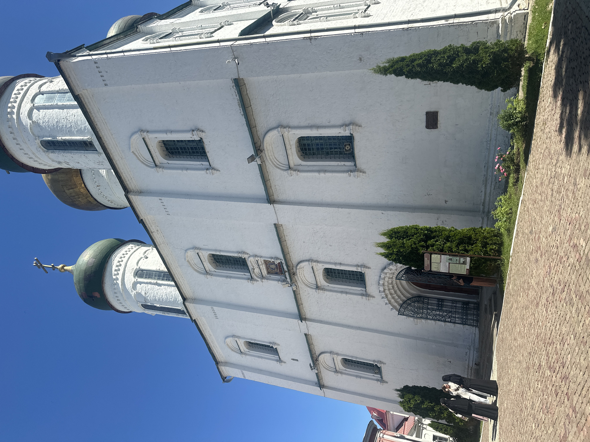 The image shows the Assumption Cathedral in Kolomna, Russia, on a sunny day. The cathedral is a large, white building with multiple domes, some of which are green and some are golden. The architecture is traditional Russian Orthodox. There are several arched windows on the side of the cathedral. In the foreground, there is a paved courtyard made of red bricks. Three people dressed in black are standing on the courtyard, facing the cathedral. One of them appears to be holding a bag. To the right of the entrance, there is a sign with text and an image. Two tall, green cypress trees are planted in front of the cathedral, one on each side of the entrance. Further to the right, another cypress tree stands in a patch of grass with some small pink flowers. The sky is clear and blue.