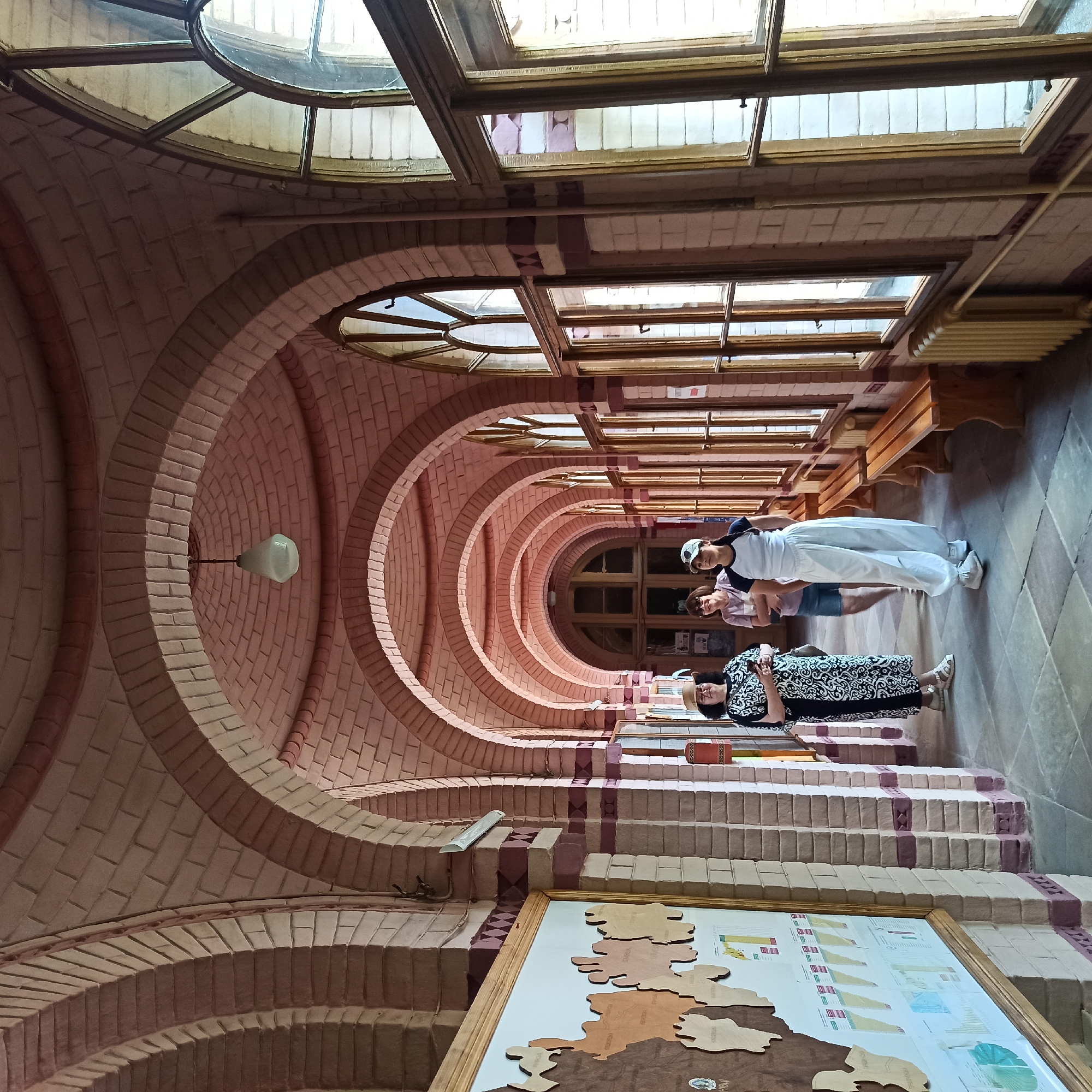The image depicts an interior view of a long, arched hallway constructed with pinkish-red bricks. The hallway is lined with wooden-framed windows on the right side and features a map displayed on a board on the left. Three individuals are present in the hallway: a woman in a patterned dress and hat stands on the left, a woman in a pink shirt and shorts is in the middle, and a young person in white athletic wear and a cap is on the right. Wooden benches are visible along the right wall. The architectural style, particularly the repeated arches and the brickwork, is characteristic of the Chernivtsi National University (formerly Residence of Bukovinian and Dalmatian Metropolitans) in Chernivtsi, Ukraine, a UNESCO World Heritage Site. The lighting appears to be natural light streaming in from the windows and an overhead lamp.
