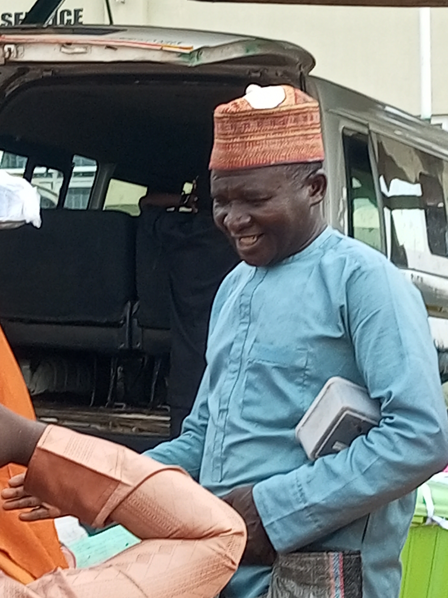 In Gombe, Nigeria, a middle-aged African man wearing a light blue traditional outfit and a patterned, reddish-brown cap is pictured outdoors. He is smiling broadly and holding a small, light-colored rectangular object in his right arm, which is tucked into his side. His left arm is bent, and his hand is visible, resting on his lap or on the clothing of another person partially visible in the frame. He is standing next to a vehicle, with the open rear door and seating area of the vehicle visible behind him. The background suggests an urban or semi-urban environment.