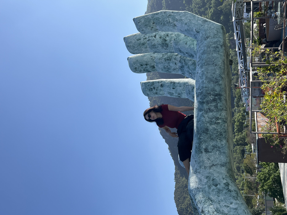 An outdoor scene captured in Nha Trang, Vietnam, features an adult female seated on a monumental, light blue-green concrete sculpture shaped like a human hand. The woman, with dark hair and sunglasses, wears a dark red short-sleeved top and dark pants, reclining on the palm portion of the sculpture while facing towards the left side of the frame. The sculpture's five fingers extend upwards and to the right. In the background, verdant, tree-covered mountains rise against a clear, bright blue sky. Below the sculpture, parts of a terraced area or rooftop garden are visible, showing various plants, railings, and metal framework structures. This location is identified as Ana Marina in Nha Trang.