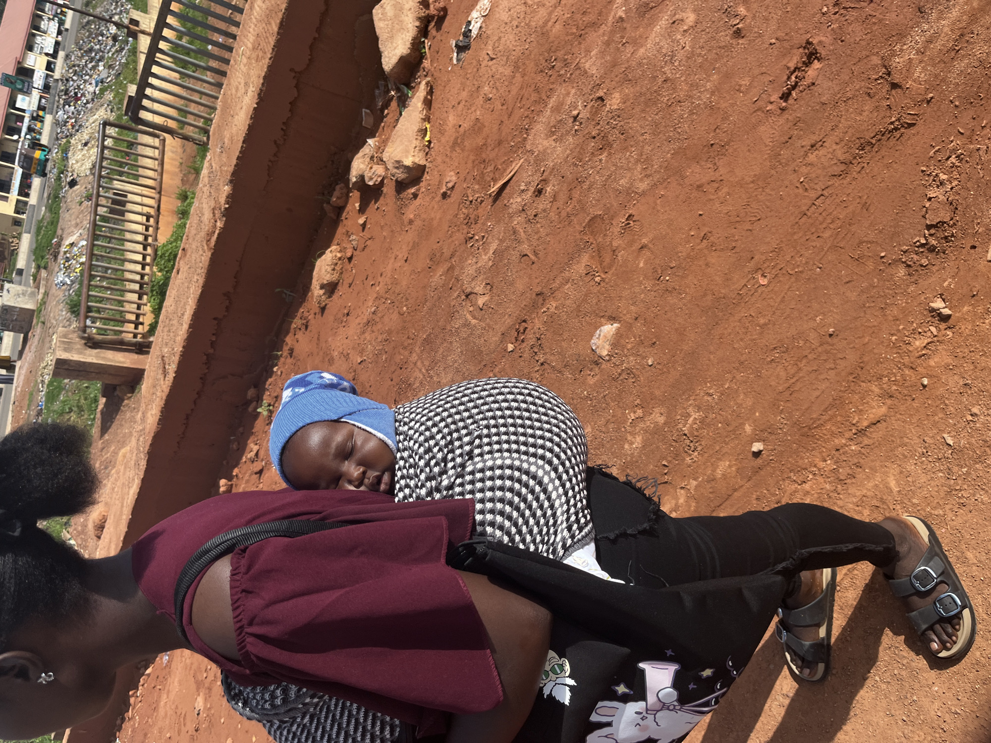 A woman carries a baby on her back in a sling. The baby is wearing a blue hat and is asleep. The woman is wearing a maroon off-the-shoulder top and black pants with a rabbit graphic. She is also wearing sandals. They are walking on a dirt path. In the background, there is a red brick wall, a metal fence, and a pile of trash. Beyond that, there are buildings and a busy street with vehicles. The scene appears to be in an urban area of Gura Top, Nigeria, characterized by its reddish-brown earth and the presence of both residential and commercial structures amidst a less-than-pristine environment.