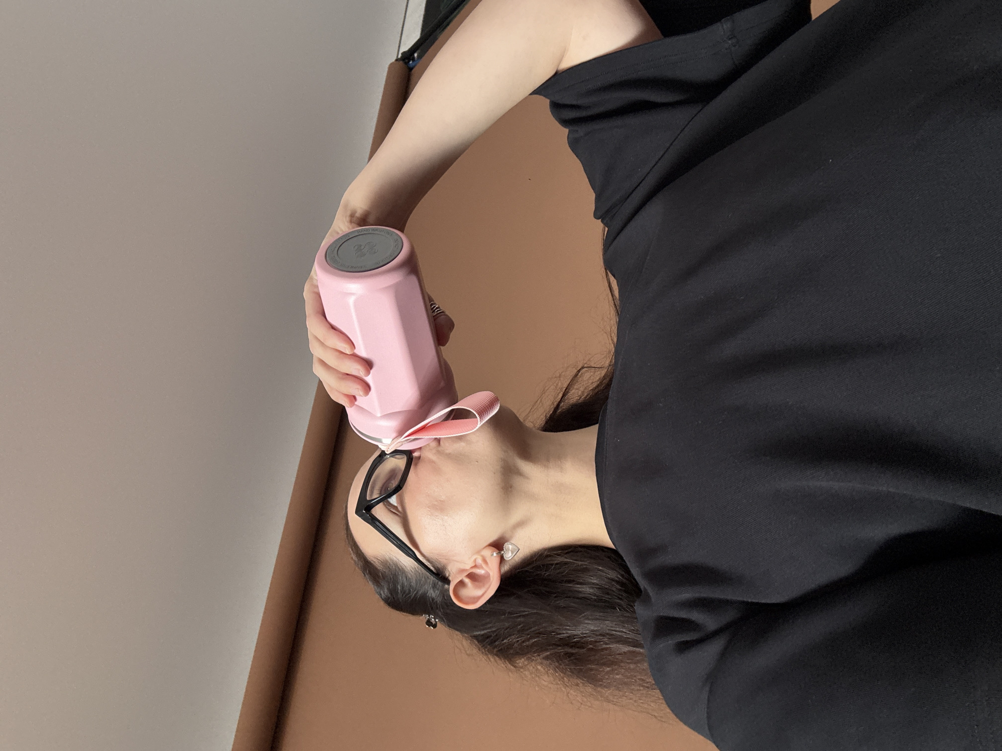 A person is drinking from a pink water bottle. They are wearing a black t-shirt and glasses. The background is a brown wall and a white ceiling. The image does not contain any information about the location in the city of Chaikovsky, Russia.