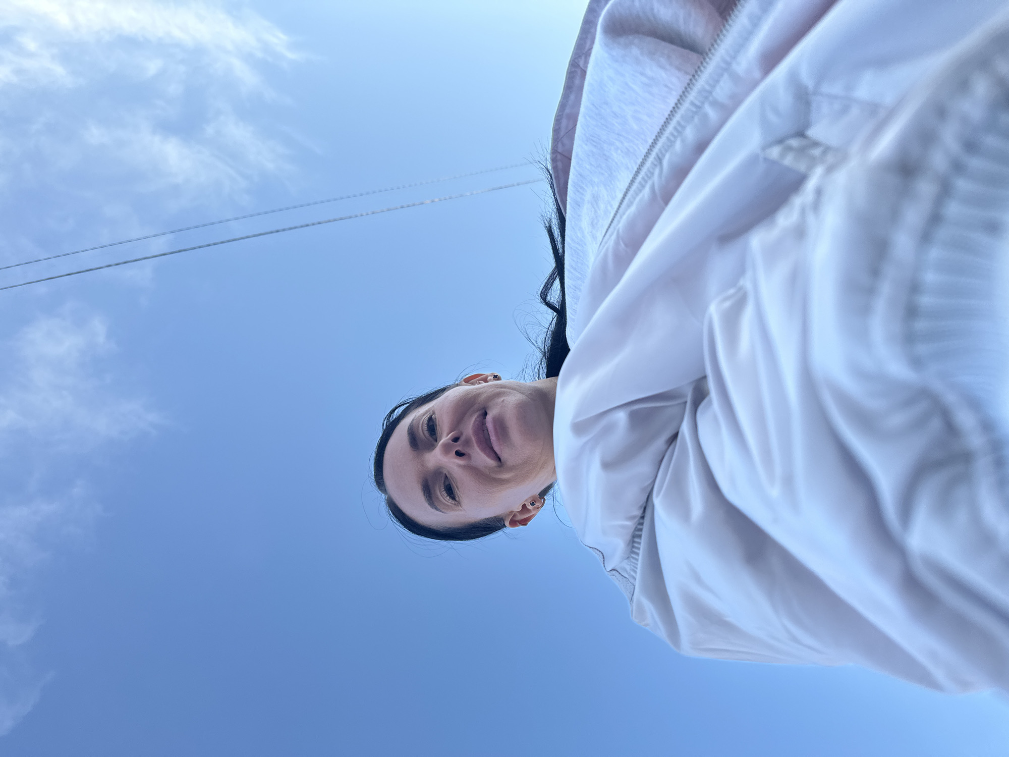 A unique low-angle perspective captures a woman looking down towards the camera against a bright blue sky. Her head and upper torso are visible in the lower half of the frame, appearing to gaze directly downwards with a calm, pleasant expression and a subtle smile. She has dark hair and wears small stud earrings. Her attire consists of a light-colored, possibly white or light pink, jacket featuring a zipper and visible fabric folds.
The upper portion of the image is dominated by a clear, vibrant blue sky dotted with sparse, wispy white clouds. Two thin, parallel lines, likely utility wires, traverse the sky in the middle ground. The bright illumination indicates it is daytime with clear weather. There is no visible text in the image. This outdoor scene is set in Ольховка, Russia.