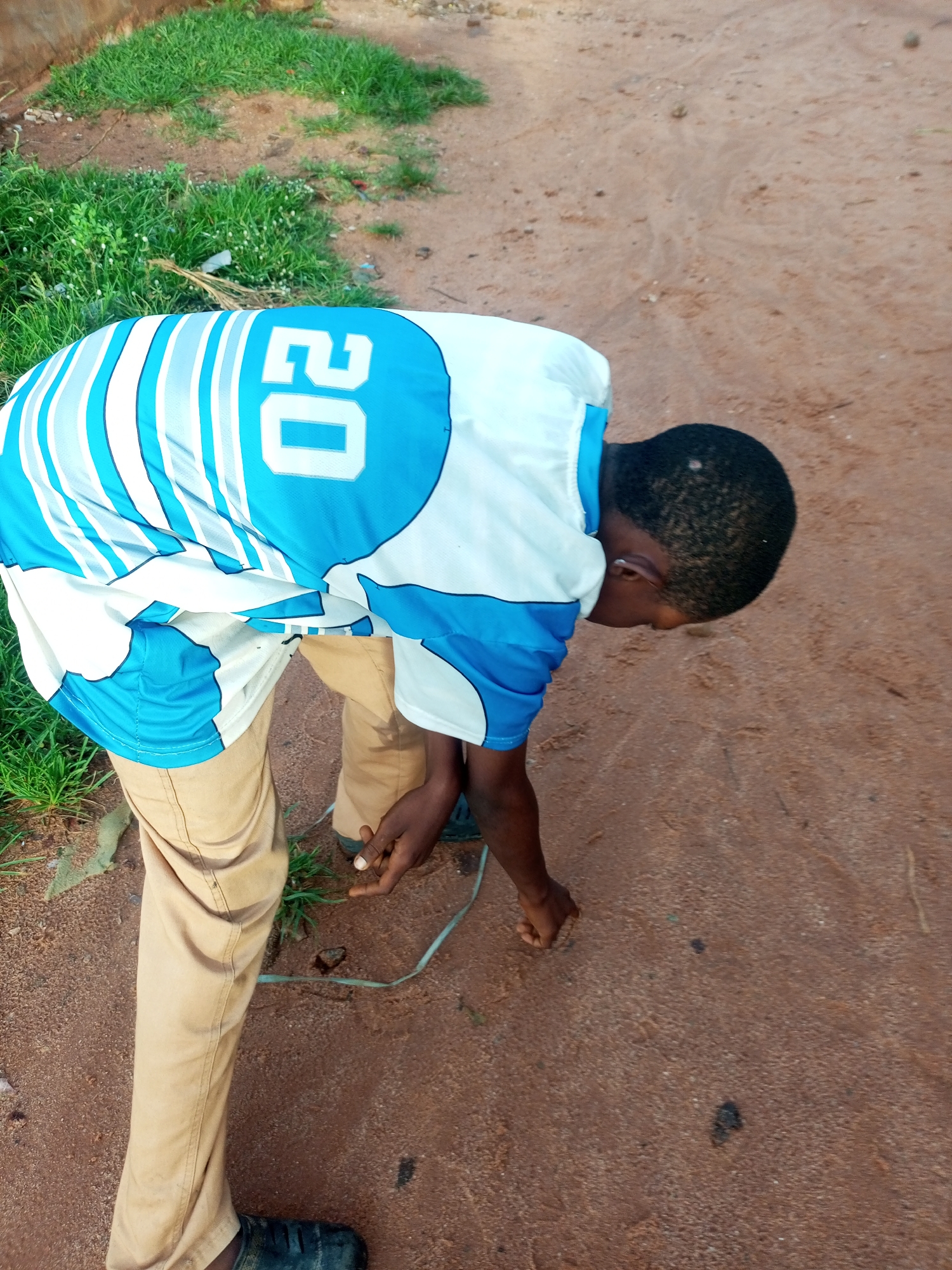 A young boy with dark skin and short, dark hair is bent over on a dirt path in Tempure, Nigeria. He is wearing a white and blue jersey with the number "02" in white on the back, and tan pants. His left hand is on the ground, appearing to interact with something on the dirt. A thin, light blue cord or string is visible near his hands and feet, extending across the path. The ground is sandy and reddish-brown, with patches of green grass to the left of the boy. The overall scene appears to be outdoors, possibly in a rural or less developed area of the city.