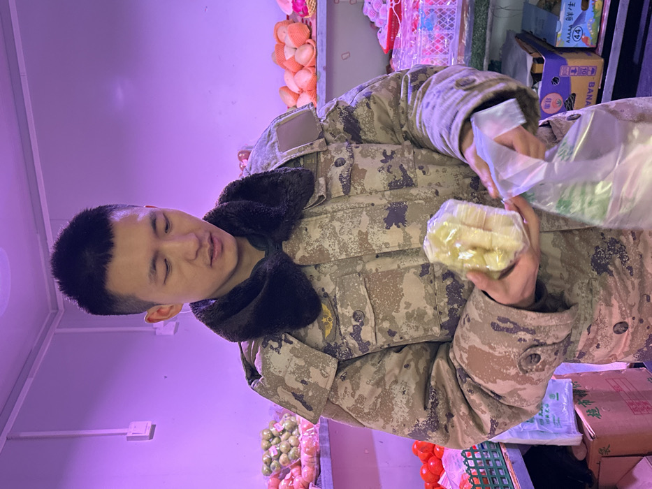 A male individual, appearing of East Asian descent, is depicted inside a retail grocery establishment in Jiangjin, China. He wears a camouflage winter jacket with a dark fur collar and a gold-colored winged star insignia on the left chest. The individual holds a clear plastic-wrapped package containing several segments of a pale-yellow fruit in his left hand and an open clear plastic bag in his right hand. His gaze is directed downwards towards these items. The background features various produce: red tomatoes in a green plastic crate, green-skinned fruits in clear plastic bags, and numerous light-colored, round fruits in mesh packaging on shelves to the right. Cardboard boxes, some labeled 'BANANA' in English and Chinese characters, are visible beneath the produce displays. The interior walls are white, and the scene is uniformly illuminated by artificial lighting, which casts a prominent purple hue. An electrical conduit and outlet are visible on the left wall.