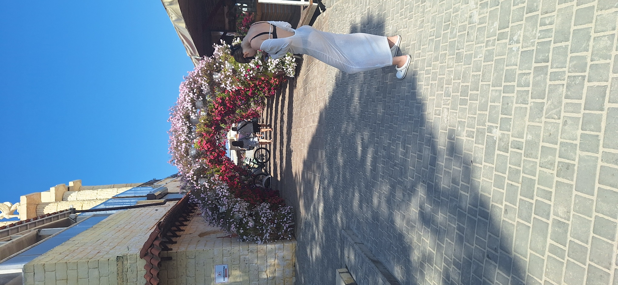 In Odesa, Ukraine, a woman in a sheer white dress and black bikini top stands on a paved walkway, looking down at her phone. She is wearing white clogs. Behind her, a floral archway made of pink and red petunias frames a pathway leading to a building. Two people are walking away from the camera through the arch. A bicycle is parked to the left of the arch. The building on the left is made of light-colored stone bricks and has a red-tiled roof. A statue of a winged creature, possibly a sphinx, sits atop columns on the building. The sky is clear and blue. The scene is illuminated by bright sunlight, casting shadows on the paved ground.