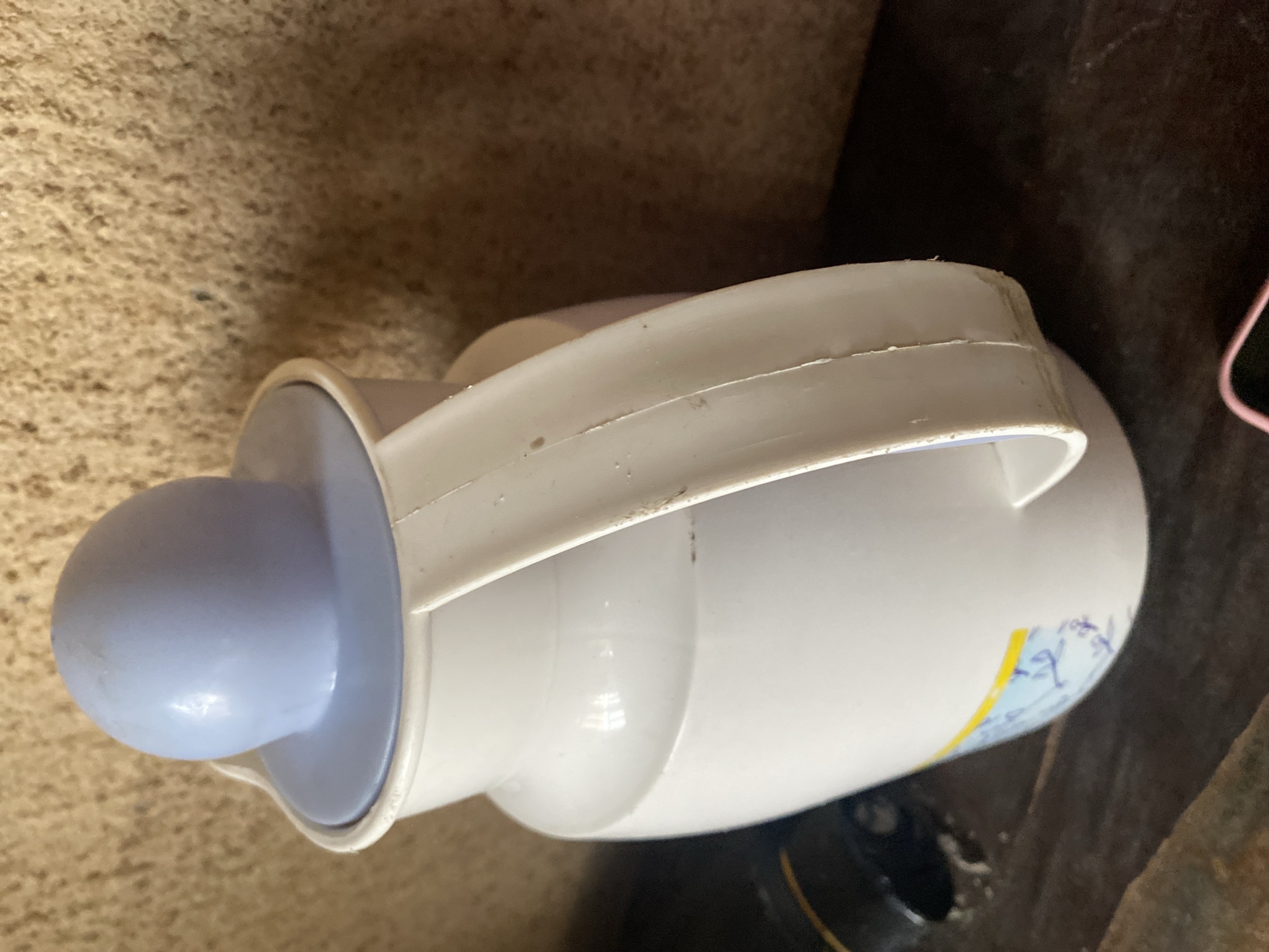 A white plastic pitcher with a light blue lid sits on a dark wooden surface. The pitcher has a curved handle on the right side and a decorative blue and yellow sticker on its lower front. The background is a textured, beige wall. The scene appears to be indoors, possibly in a home in Jauro Yinu, Nigeria.
