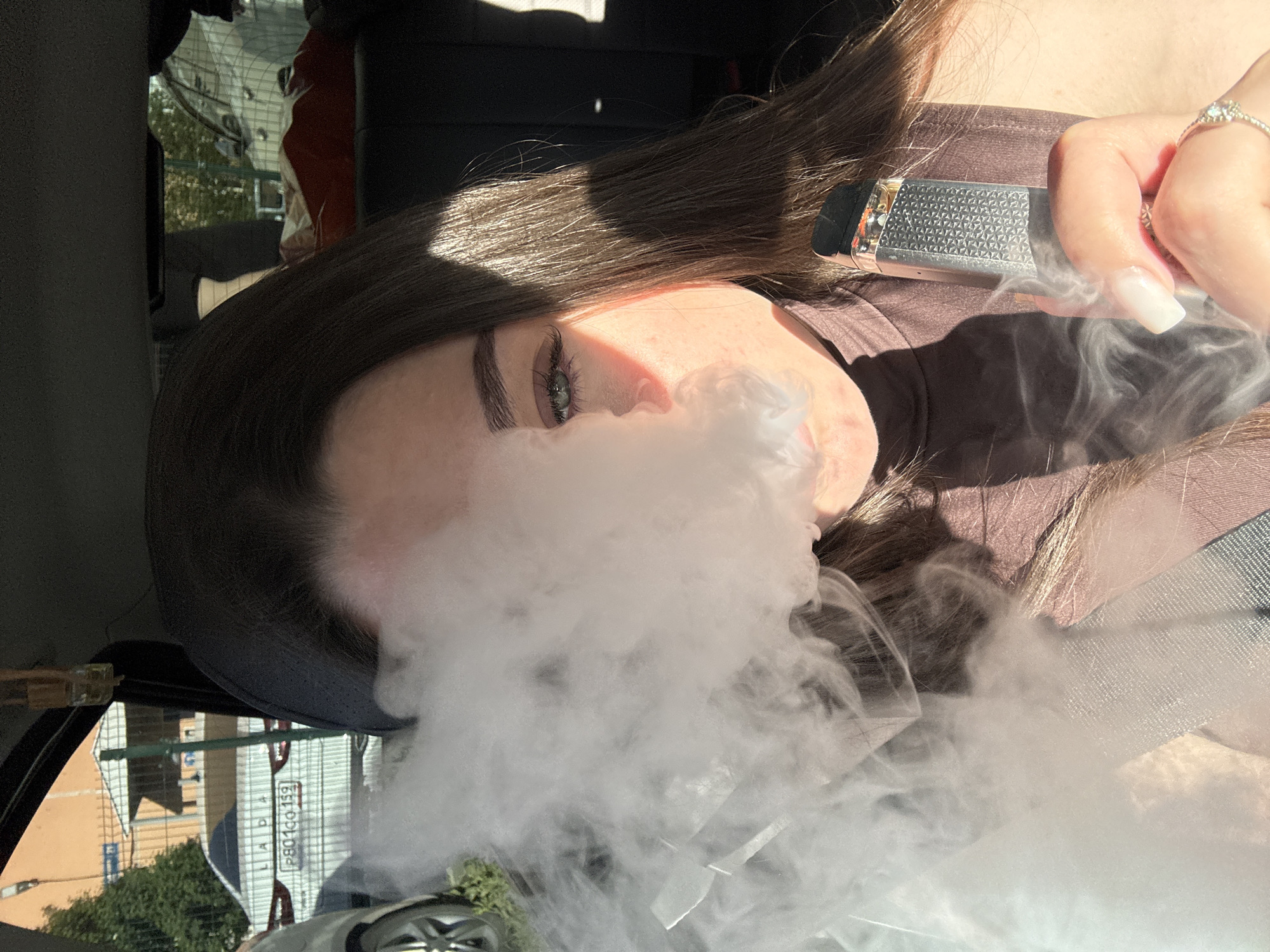 A young woman is visible from the chest up, inside a car. She is exhaling a large cloud of vapor from what appears to be a vape pen held in her right hand. The vapor obscures much of her face. She has long, dark hair and light-colored skin, and is wearing a brown top. The car is likely located in Perm, Russia, as a license plate visible through the car window reads "P801CO 159," where 159 is the code for the Perm Krai region. The photograph appears to have been taken in daylight.