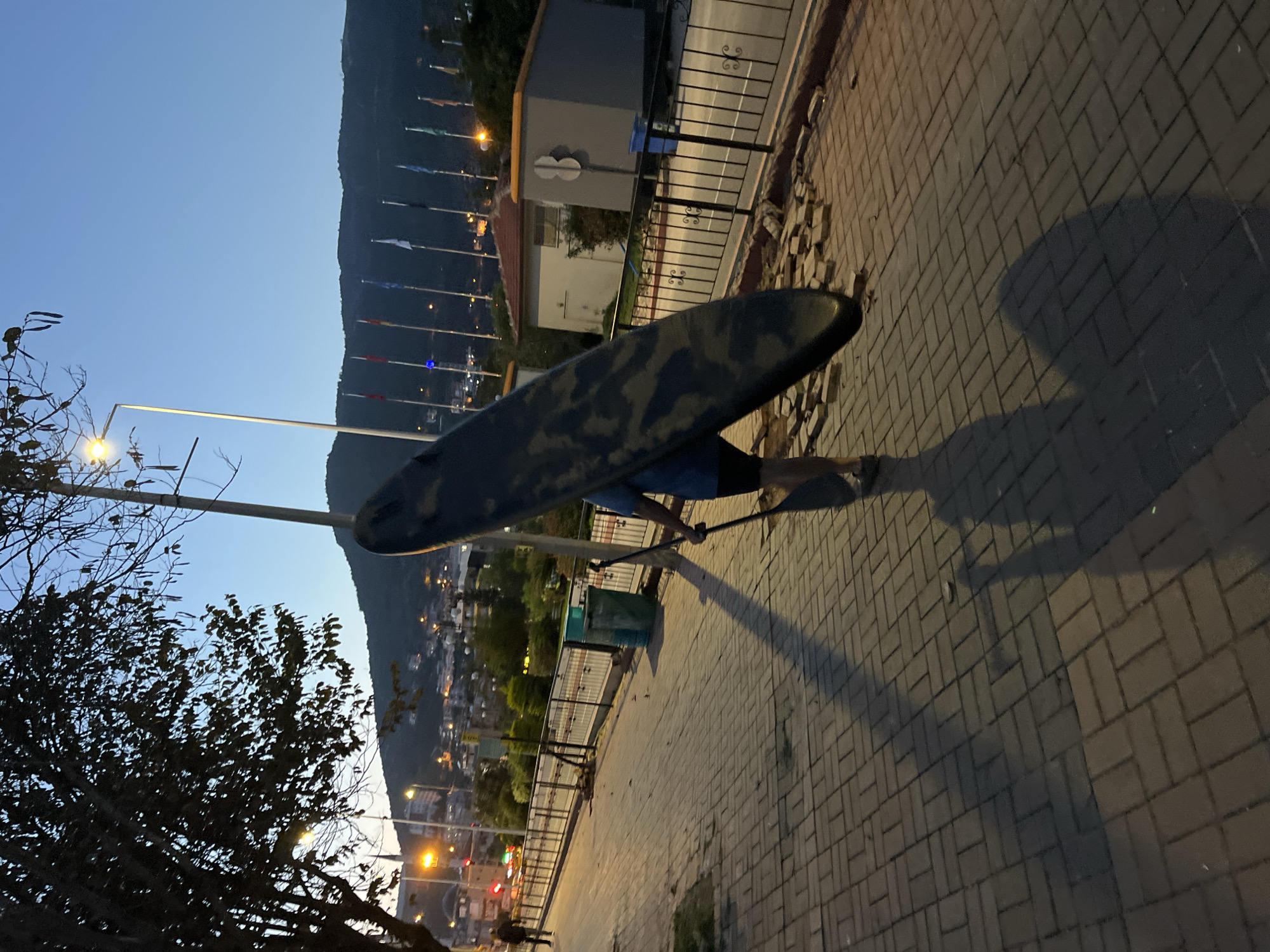 A side-angle, low-light photograph depicts a person carrying a camouflage-patterned paddleboard on a paved brick walkway in Alanya, Turkey. The individual, wearing a blue shirt and dark shorts, balances the board on their head. A long shadow is cast on the walkway. A long-handled paddle is visible in their hand.
The walkway is bordered by a black metal railing and a green trash receptacle. Behind the railing is a view of the city and a distant mountain range, with several flags flying along the street. Streetlights and buildings are visible, contributing to the ambient light. To the left is a tree. The sky is light. The scene suggests it is dusk or early evening.