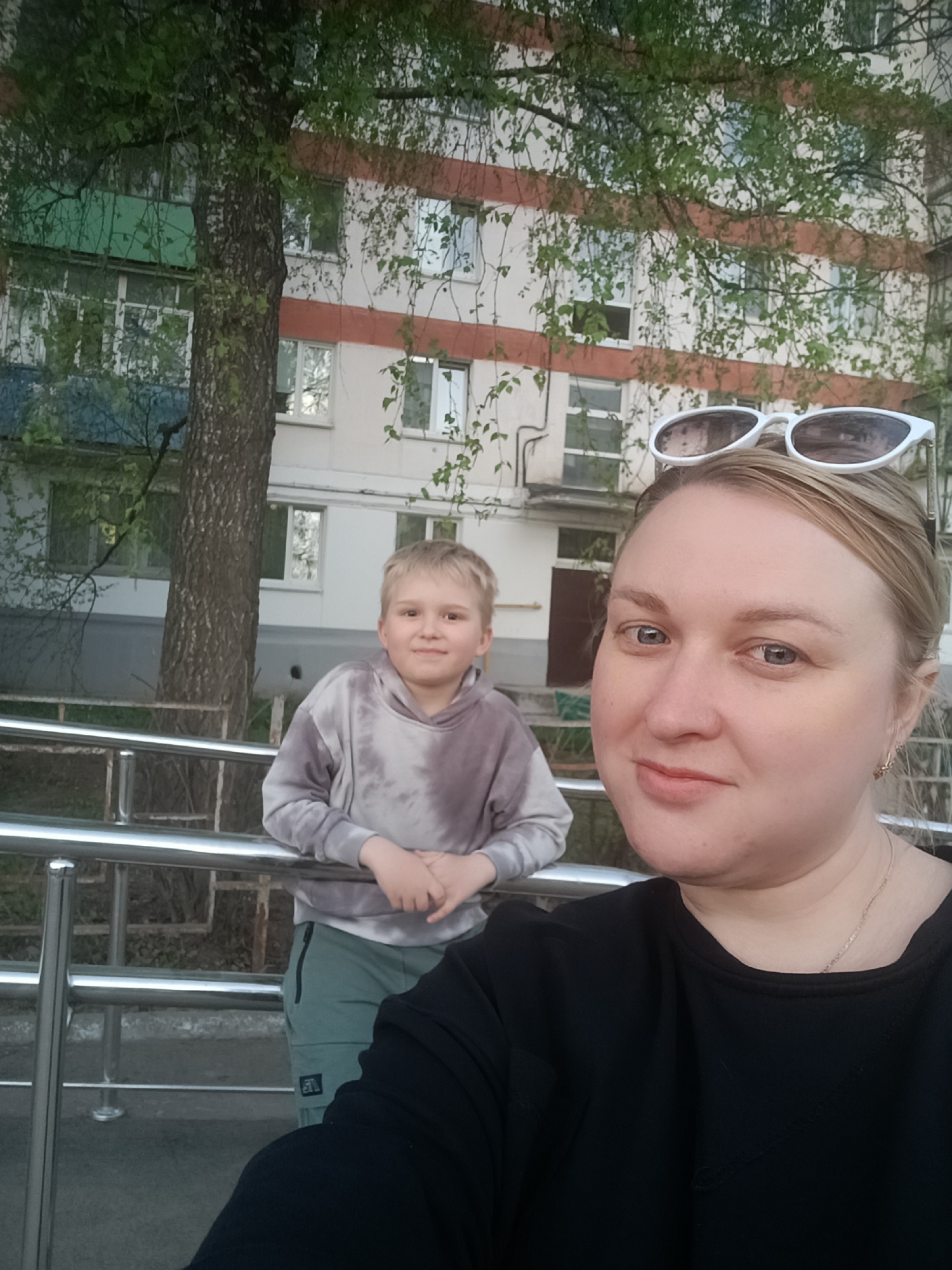 The image shows a woman and a boy in front of a building. The woman is in the foreground, wearing a black shirt and sunglasses on her head. The boy is behind her, leaning on a metal railing. He is wearing a tie-dye hoodie and green pants. Behind them is a building with multiple windows and some trees with green leaves. The location is the city of Chaikovsky, Russia.