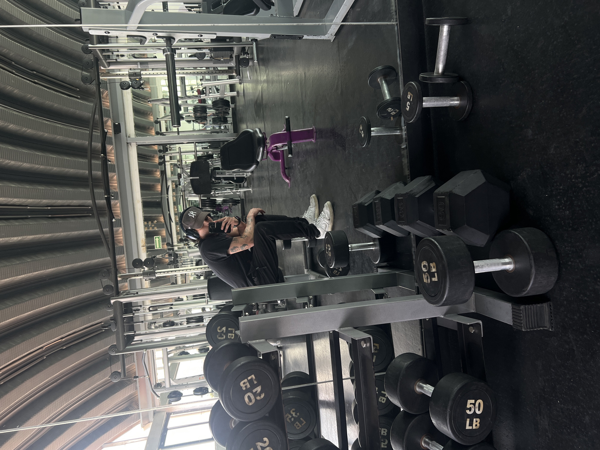 The image captures an indoor gym environment, with a person seated on the floor in the foreground. The individual, dressed in a black t-shirt, black pants, and white sneakers, wears a dark baseball cap with a white "NY" logo and headphones. Tattoos are visible on their arms as they hold a smartphone, seemingly taking a selfie or recording.
The gym features a prominent black dumbbell rack in front of the person, displaying various dumbbells labeled "50 LB", "32 LB", and "20 LB". Another "5 LB" dumbbell is visible further to the right. A purple exercise bench stands out in the mid-ground. The floor is dark and appears to be rubberized. In the background, cable machines and other workout equipment are visible, with reflections in large wall mirrors. The ceiling has a distinct corrugated metal structure, and natural light enters through windows, indicating it is daytime. The scene suggests a moment of rest or reflection during a workout session.