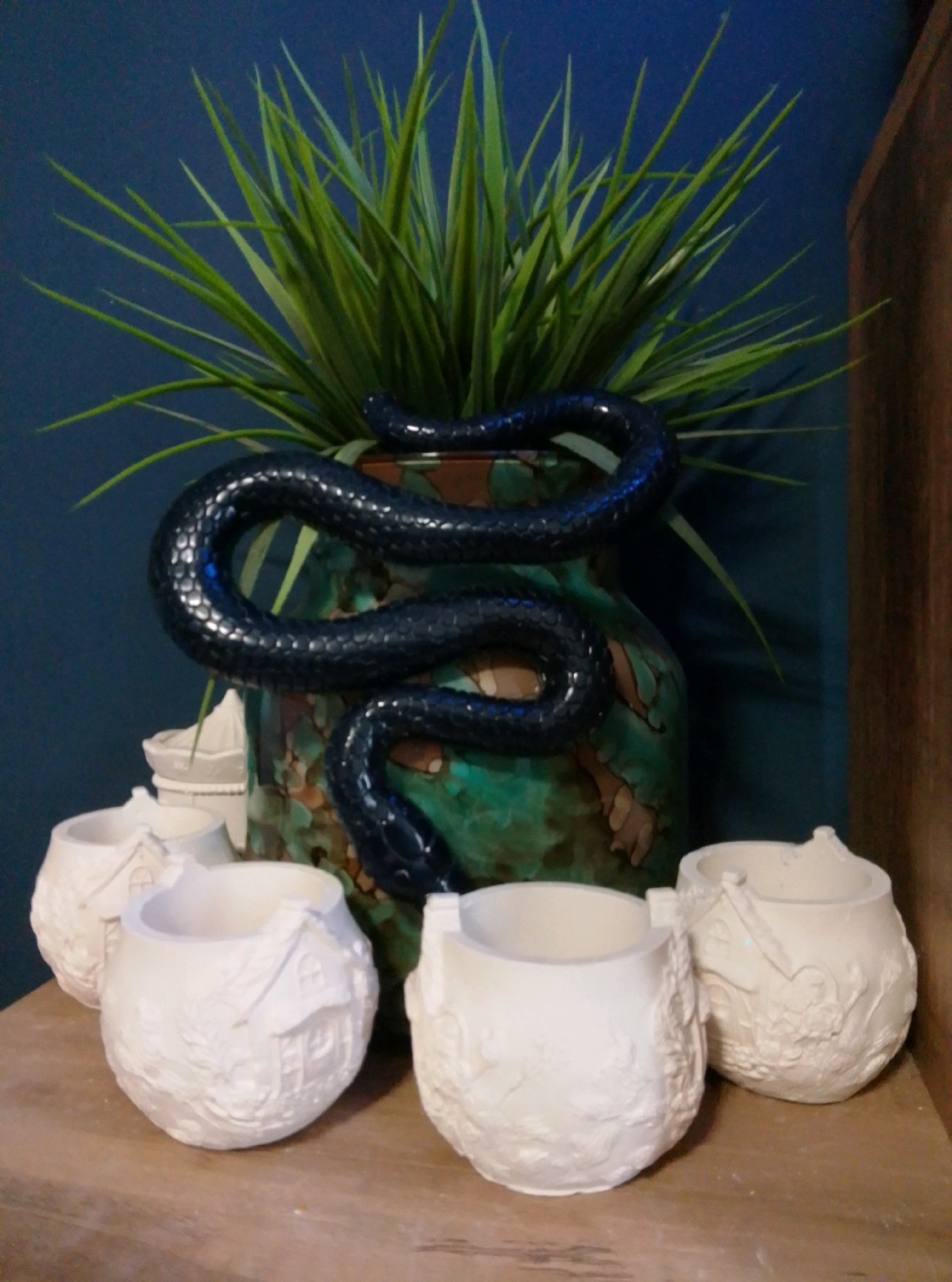 In a domestic setting, likely a shelf in a home in Wedel, Germany, a collection of decorative objects is displayed against a dark blue wall. Dominating the center is a large, emerald green vase with a mottled brown and tan pattern. Coiled around the neck and upper body of the vase is a black, sculpted snake with a textured, scaled surface and a glossy finish. Emerging from the top of the vase is a dense clump of artificial green grass, its spiky blades fanning outwards. In front of the vase, arranged on a wooden surface, are three small, unglazed white ceramic pots. Each pot is spherical and features a raised, intricate relief design depicting a quaint cottage with a thatched roof, windows, and surrounding foliage. The pot on the left is partially obscured by the one in the center. The pot on the right is the most prominent of the three, its details clearly visible. The overall scene suggests a collection of decorative, possibly handcrafted, items.