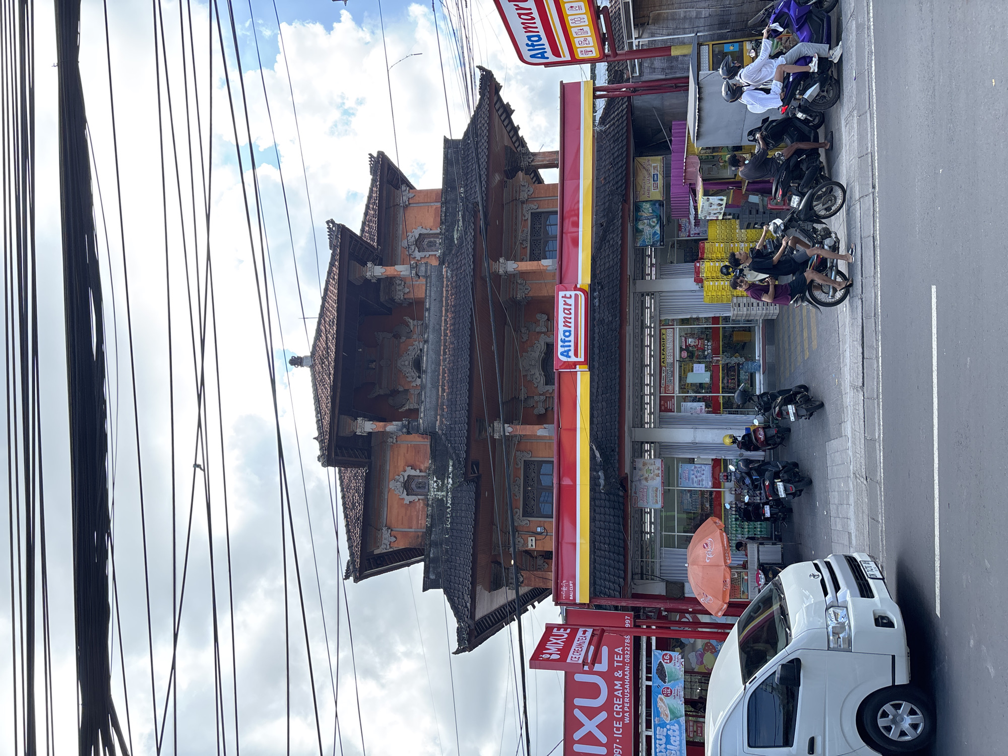 A daytime street scene in Kuta Selatan, Indonesia, features a blend of traditional architecture and modern commerce under a bright, cloudy sky. A dense network of overhead power lines spans across the upper left portion of the image.
On the right, a multi-story building with distinct traditional Balinese architecture is visible, characterized by its ornate carvings, decorative elements, and a multi-tiered, dark-tiled roof. Directly next to it is an Alfamart convenience store, identifiable by its prominent red and white signage.
On the paved street, a white van is parked. The sidewalk in front of the Alfamart is occupied by several parked motorbikes, on which multiple individuals are seated, appearing to be resting or waiting. Some wear helmets, while others do not. An orange umbrella is also present on the sidewalk.
Visible text includes "Alfamart" (on multiple signs), "MIXUE ICE CREAM & TEA," "997 ICE CREAM & TEA," a WhatsApp number "WA PERUSAHAAN: 0822788 997," "BALI CLIFT," "ALFAGIFT," and "DI SINI." Various product displays and additional Indonesian text on banners are partially discernible inside and around the stores. The scene depicts a typical, calm moment of daily activity.