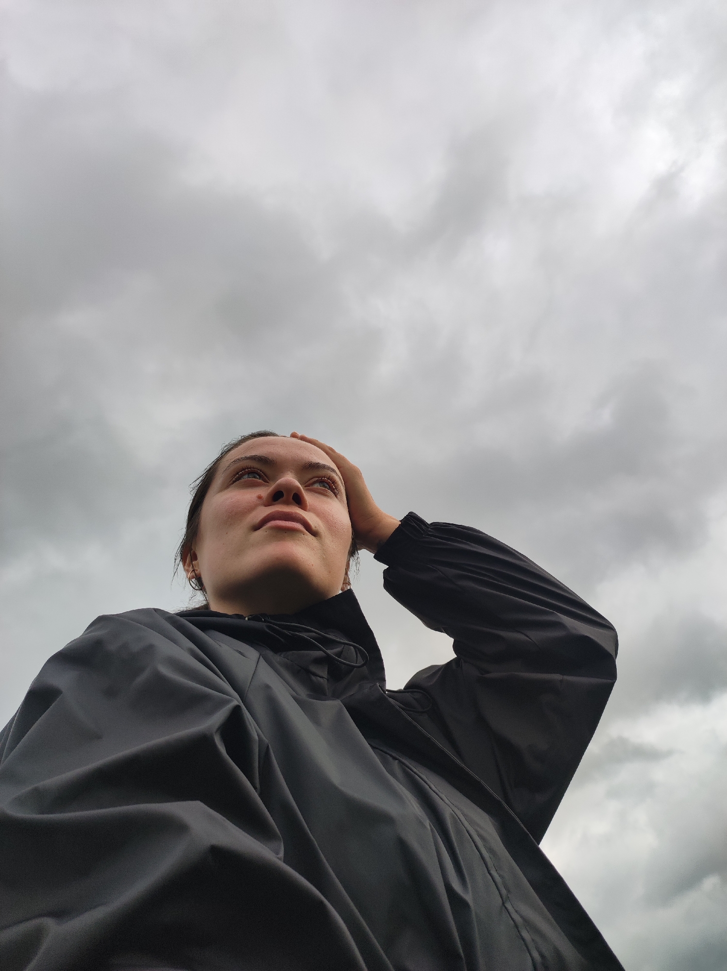 This low-angle, medium close-up shot features a young person, likely female, looking upwards against an overcast sky. The subject, positioned in the lower left and central portion of the frame, has a contemplative and serene expression, with her gaze directed towards the cloud-filled sky.
She is wearing a dark, likely black or dark grey, waterproof jacket or raincoat, which covers her torso and arms. The fabric of the jacket appears textured and somewhat reflective in the diffused light. Her right hand is raised, with her fingers gently resting near her forehead or hairline, a gesture that could indicate shielding her eyes, deep thought, or a moment of reflection. A small, simple hoop earring is visible in her left ear.
The background is dominated by an expanse of grey, cloudy sky, with varying tones of white and dark grey suggesting dense cloud cover and an absence of direct sunlight. The lighting is soft and diffused, characteristic of an overcast day. The overall mood of the image is one of quiet observation or introspection. No specific location cues are visible within the image itself beyond the sky, but the context indicates the setting is Фоки, Russia. There is no visible text.