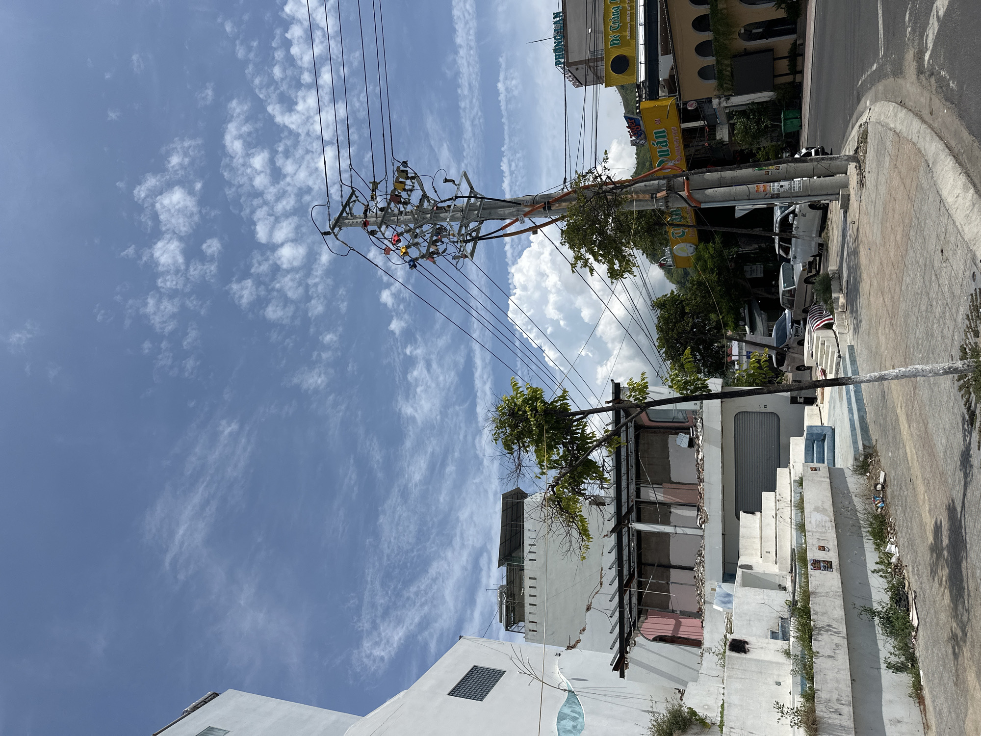 The image captures a street scene in Bac Nha Trang Ward, Vietnam, under a partly cloudy blue sky. A white building with a distinctive blue wave design on its lower exterior is on the left. Adjacent to it, another building shows signs of disrepair, with exposed beams and unfinished construction. A small, bare-branched tree stands in front of this building.
A prominent utility pole with numerous wires and colorful insulators dominates the center of the frame. The road curves to the right, with several vehicles parked along the sidewalk, including SUVs and sedans. Commercial establishments are visible in the background, with signs in Vietnamese. One sign features a yellow background with the text "Tiger" and "Quan 01". Another building has an arched facade and a dark sign. The sidewalk is paved with concrete tiles, and debris is scattered near the curb. A large, white cumulus cloud is visible in the distance, partially obscured by the utility pole and buildings. The overall impression is of a partially developed or under-renovation urban environment.