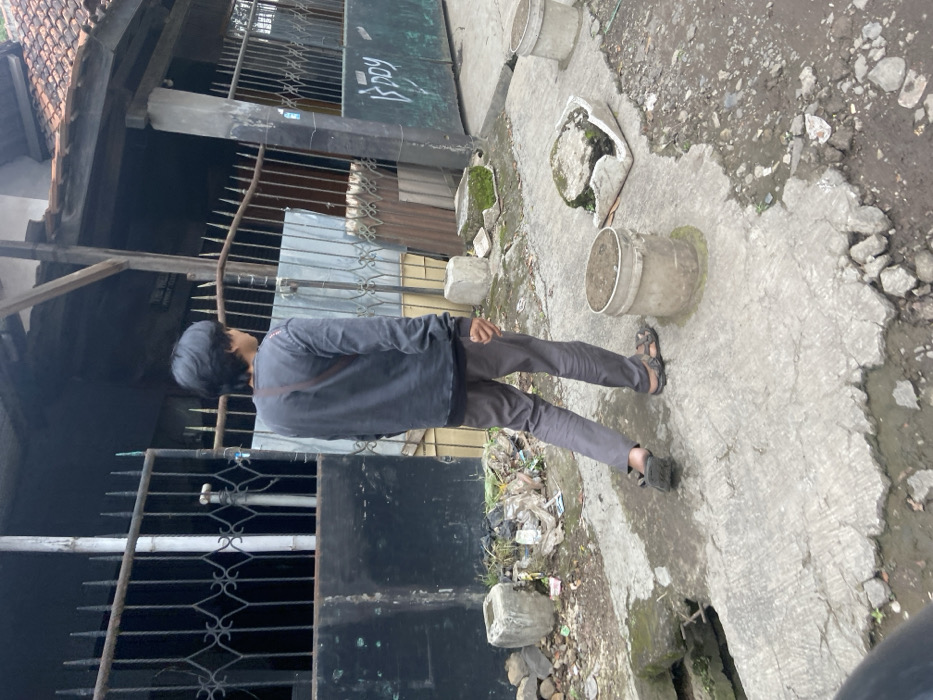 The image shows a young man walking away from the camera on a cracked concrete sidewalk. He is wearing a dark grey long-sleeved shirt and brown pants. The man is wearing black sandals. He is walking past a house with a black metal gate and a broken concrete drain. There is a metal bucket on the right side of the image. The location is on a street in Baleendah, Indonesia at the coordinates -7.021, 107.673.