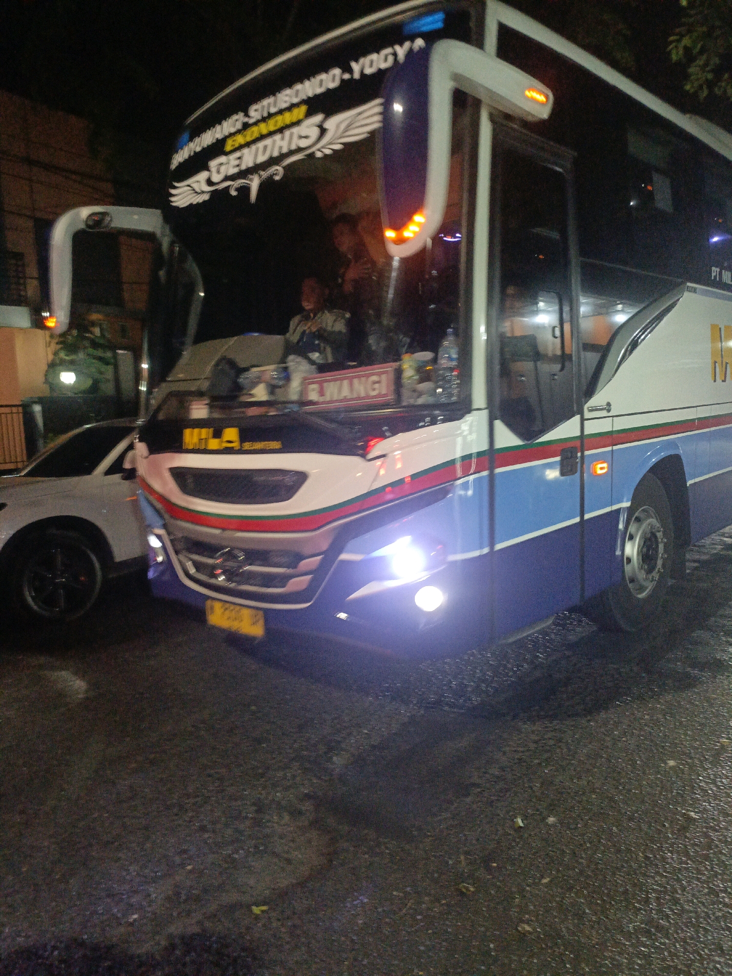 An eye-level, slightly low-angle outdoor medium shot captures a blue and white intercity bus, identified by the text "EKONOMI GENDHIS" and "BAYUWANGI SITUBONDO-YOGYA" on its upper front. The bus is parked on a wet asphalt road at night, illuminated by its own headlights and streetlights, which cast a soft glow on the surroundings. A white SUV is parked to the left of the bus. The bus's windshield is visible, with indistinct figures inside. The environment appears to be a dimly lit street, possibly in a commercial or residential area, with some foliage visible in the background. The overall atmosphere is calm and still.