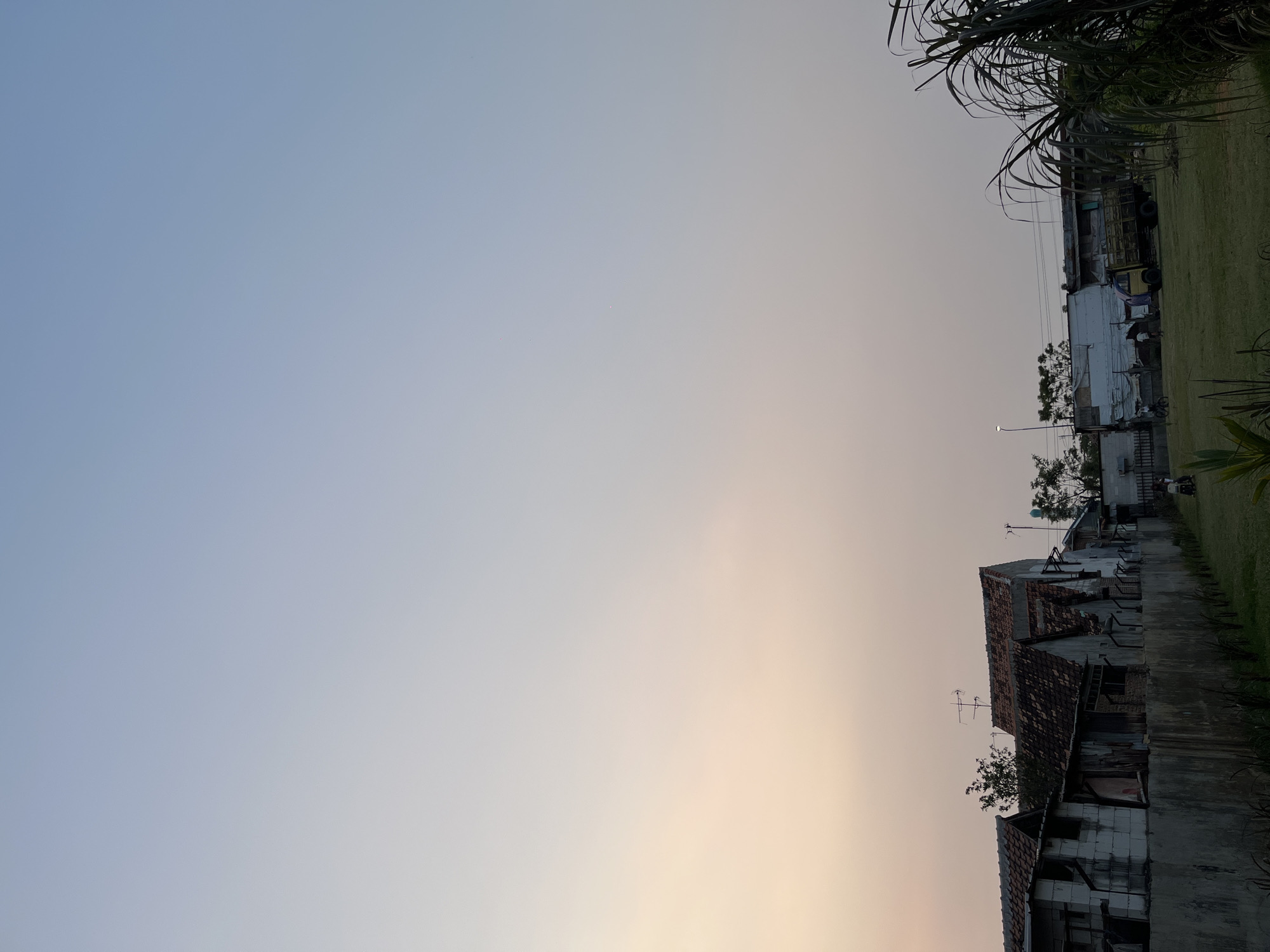 The image depicts a street scene in Pekalongan, Indonesia, during what appears to be either early morning or late afternoon, given the soft, gradient lighting of the sky. In the foreground, a grassy area is visible with some sparse vegetation and tall, reedy plants on the right. A concrete wall runs horizontally across the middle ground, behind which a row of buildings with tiled roofs is situated. These buildings appear to be a mix of residential and perhaps small commercial structures, some showing signs of age or disrepair. A yellow truck is parked to the right of these buildings, and several indistinct figures are visible near it and on bicycles, suggesting some human activity. A street lamp stands further back in the scene. The sky transitions from a light orange or peach hue near the horizon to a pale blue overhead. The overall impression is one of a quiet, everyday moment in a semi-urban or peri-urban environment.