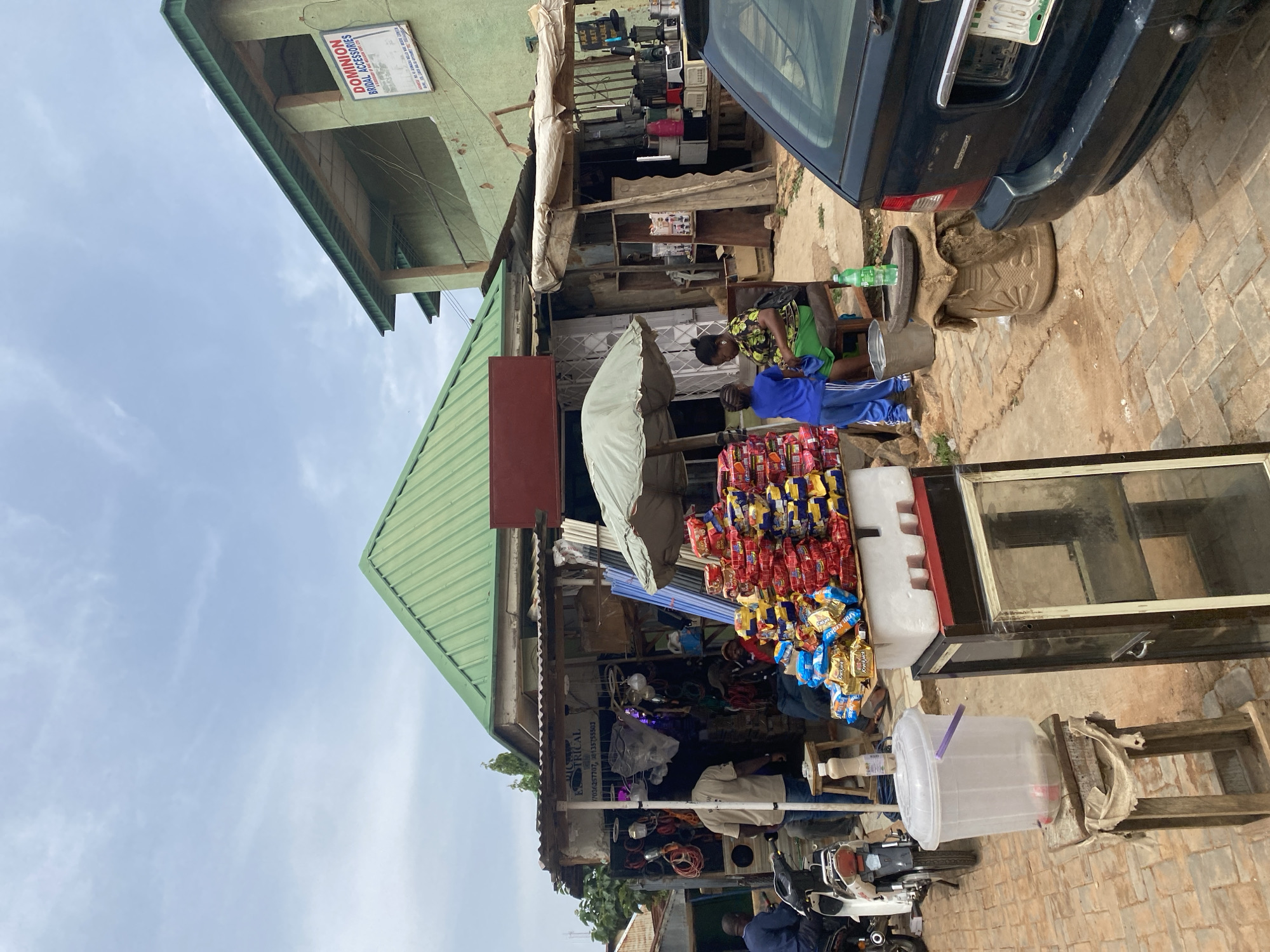 This image depicts a street scene in Jos, Nigeria, likely in a market area. The foreground features a display of packaged snacks, predominantly colorful bags of chips, stacked high on a white foam board atop a glass-fronted cabinet. To the left, a large clear plastic bucket with a purple handle sits on a wooden stool, with a bottle on its side inside. Further left, a white motorcycle is parked.
In the middle ground, two women and a child are visible. One woman, wearing a green and black patterned top and a green skirt, sits on a chair. Beside her, a child in a blue tracksuit stands. Behind them, an electrical shop is apparent, with wires, light fixtures, and plumbing supplies displayed outside. A man in a tan shirt stands near the entrance of the electrical shop.
The background reveals a green-roofed building with a sign that reads "DOMINION BRIDAL ACCESSORIES." Another building with a weathered green facade is adjacent. The overall environment appears to be an outdoor market or commercial street with stalls and shops. A dark-colored car is parked on the right side of the image. The ground is paved with interlocking bricks and dirt. The sky is overcast with scattered clouds.