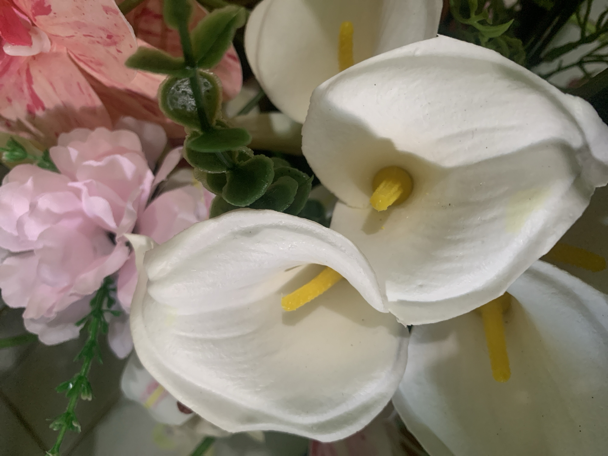 This is a close-up, eye-level, vertical shot of a bouquet of artificial flowers. The primary subjects are three white calla lilies with bright yellow spadices. There are also hints of pink and white peony-like flowers and green foliage in the background, suggesting an indoor setting. The lighting is bright, creating highlights on the smooth surfaces of the calla lilies. The image does not depict any people, actions, or interactions, and there is no contextual information provided to place it within Kuala Pembuang, Indonesia.