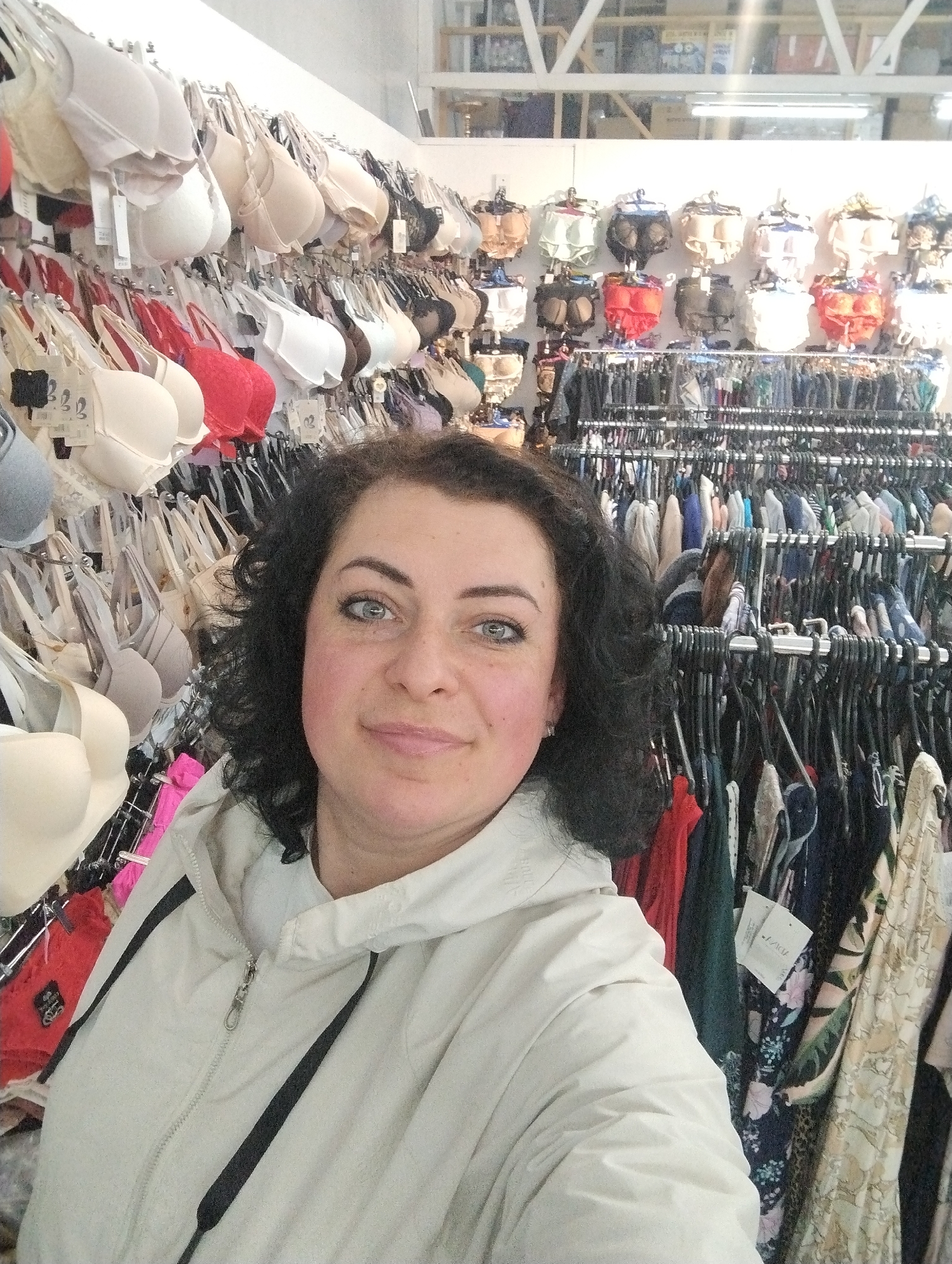 The image depicts a woman taking a selfie in a clothing store. She has dark curly hair, fair skin, and light-colored eyes. She is wearing a light-colored zip-up jacket with black drawstrings. Behind her, there are racks of clothing. One rack is filled with bras in various colors and styles. Another rack contains hanging dresses and other garments. The background suggests a retail environment with shelving and additional merchandise visible. The location is the city of Votkinsk, Russia.
