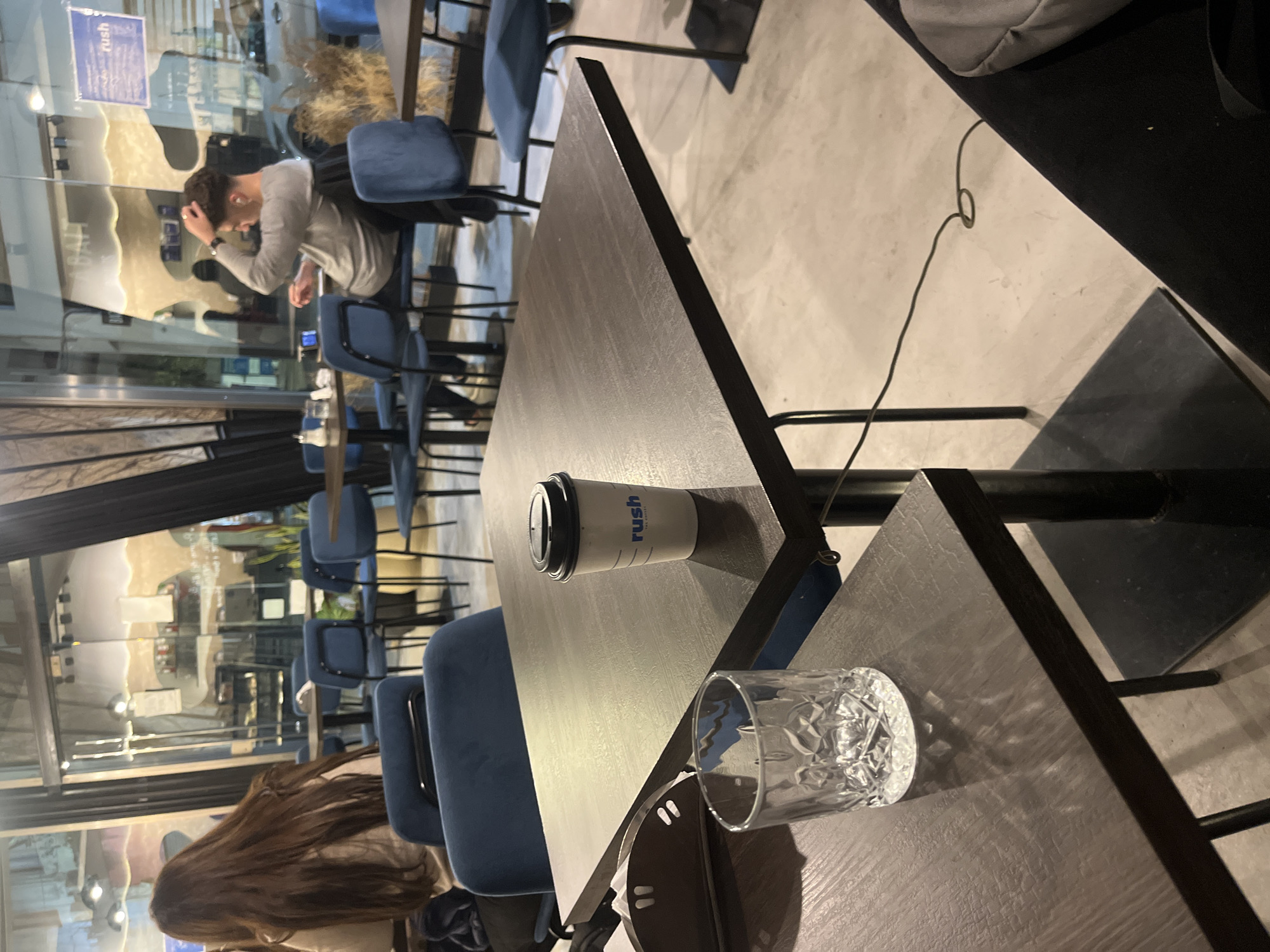 This media file captures an indoor scene within a cafe or restaurant, likely in Buenos Aires, Argentina, characterized by its modern and slightly industrial aesthetic. The perspective is from a table, looking across the establishment through large glass windows.
**Subjects:** Two individuals are visible. In the mid-ground, reflected in the window, a person with short, dark hair, wearing a light-colored long-sleeve shirt, is seated at a table, appearing to look down or contemplate something, with a hand raised towards their head. In the foreground, a person with long, dark brown hair is seated at a table, facing away from the camera. The space appears moderately occupied but not crowded.
**Objects:** The foreground features a dark wooden table where a white paper cup with a black lid, bearing the text "rush," and a clear, textured glass are placed. Other dark wooden tables with blue upholstered chairs, supported by black metal frames, are scattered throughout the venue. A black cable is visible on the light-colored floor in the upper right. The large windows reflect interior elements, including other tables, chairs, and what appear to be stylized white, wavy wall panels or decor. Some dried, bushy plants are also visible in the background, adding to the decor. A gray bag is partially visible on a seat in the upper right corner.
**Activity/Event:** People are seated at tables, suggesting a casual dining or cafe experience, possibly enjoying drinks or waiting for service. The person in the background seems engaged in a personal moment.
**Notable Details:** The scene takes place indoors. The lighting indicates it is likely evening or nighttime, with artificial lights illuminating the interior and creating prominent reflections on the large glass windows. The "rush" logo on the coffee cup is also visible as a reflection on the window. The overall ambiance appears calm and contemporary.