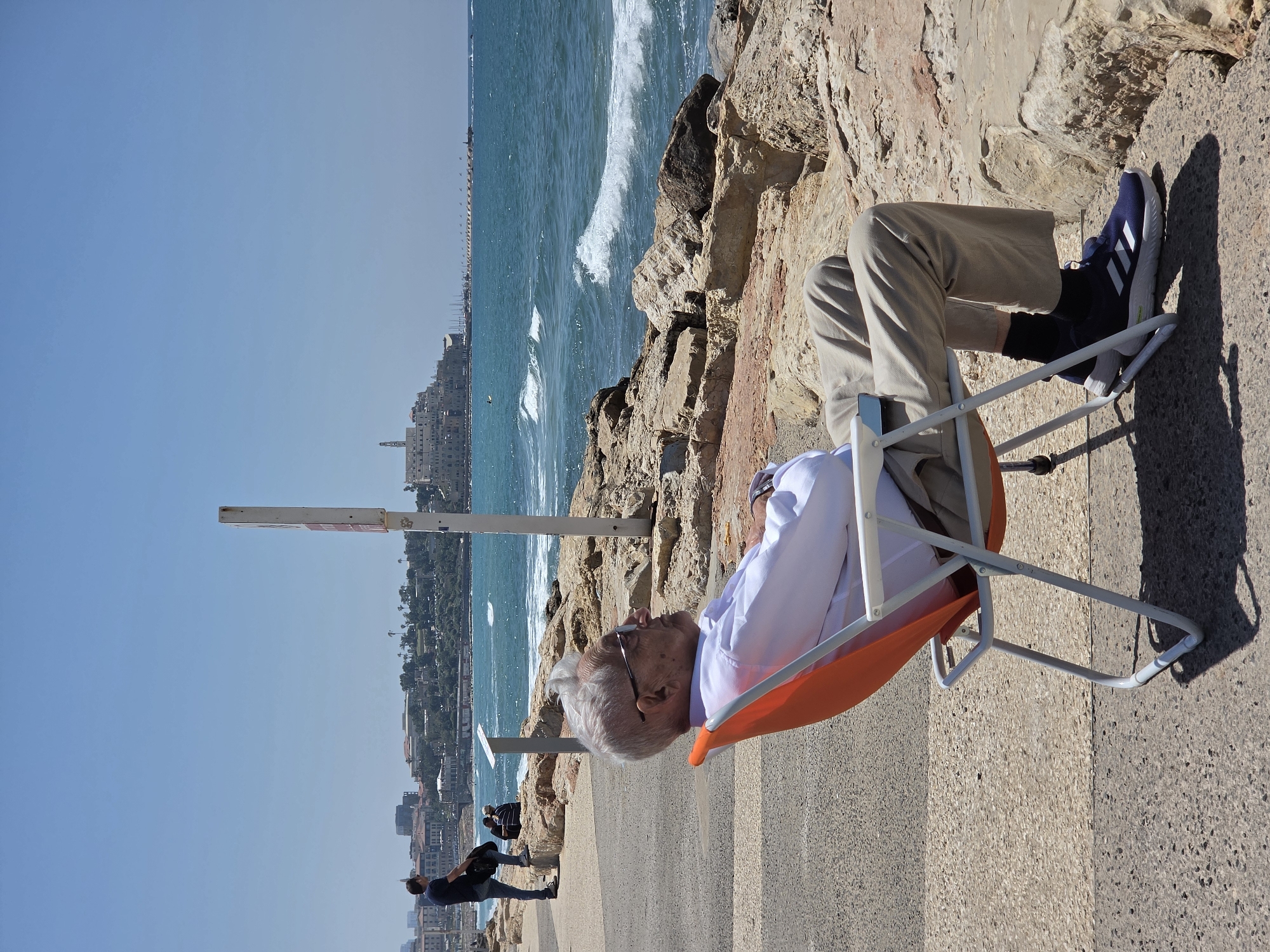 An elderly man with white hair and glasses is relaxing in an orange and white folding chair on a concrete pathway. He is wearing a white collared shirt, light-colored trousers, and dark blue sneakers. His legs are crossed, and he appears to be resting or observing the view.
The scene is a sunny coastal area in Tel-Aviv, Israel, with the Mediterranean Sea stretching out behind the man. Moderate waves are visible breaking against the rocky shore. In the distance, a cityscape with numerous buildings and some greenery lines the coastline under a clear blue sky. Two other people are visible walking further down the pathway in the distance.
The bright sunlight indicates a clear day, likely mid-morning or afternoon, casting distinct shadows from the man and his chair. A white signpost with a red rectangular top is positioned behind the man, but any text or symbols on it are illegible. The rocks along the shore exhibit various shades of brown and tan. The overall atmosphere appears peaceful and calm.