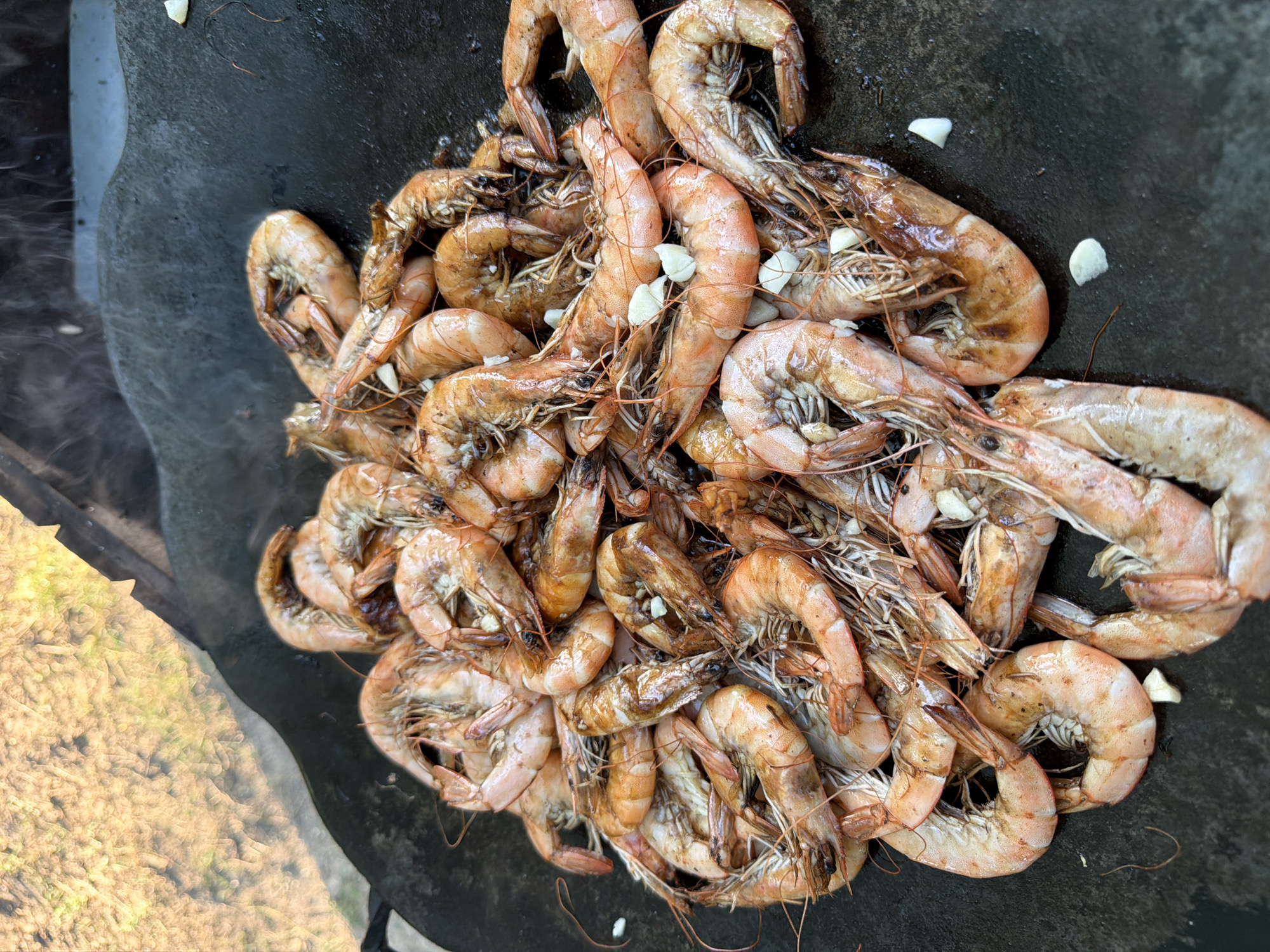 A top-down, close-up shot shows a large pile of cooked shrimp on a dark, circular cooking surface, likely a griddle or a large skillet, outdoors. The shrimp are various shades of orange and pink, with some showing char marks from cooking. Several pieces of chopped garlic are scattered amongst the shrimp. Wisps of steam are visible rising from the shrimp, indicating they are still hot. The background is out of focus, showing a blurry, natural environment with hints of green and brown, possibly a garden or grassy area. The overall impression is of a meal being prepared or served outdoors. The location in Vatra, Moldova, is not discernible from the visual information in the image.