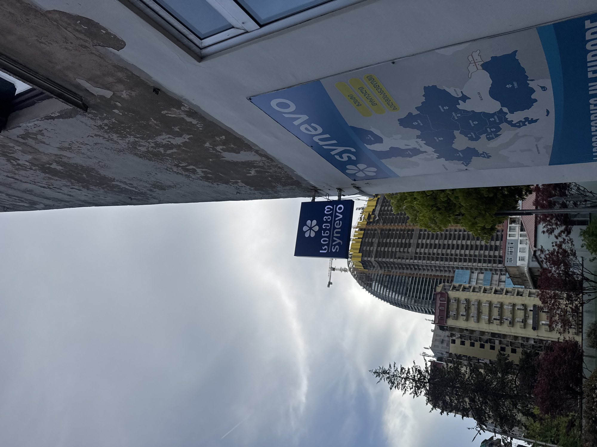Here's a description of the image:
The image appears to be taken from a low angle, looking up at the underside of a building overhang. Visible are signs for "Synevo" in both English and Georgian script, alongside a map of Europe showing marked locations.
In the background, a cloudy sky dominates the view. Below the sky, we can see urban buildings, including a modern high-rise under construction, and other older multi-story buildings with visible windows and balconies. Green trees are interspersed amongst the buildings. A construction crane is visible near the high-rise.