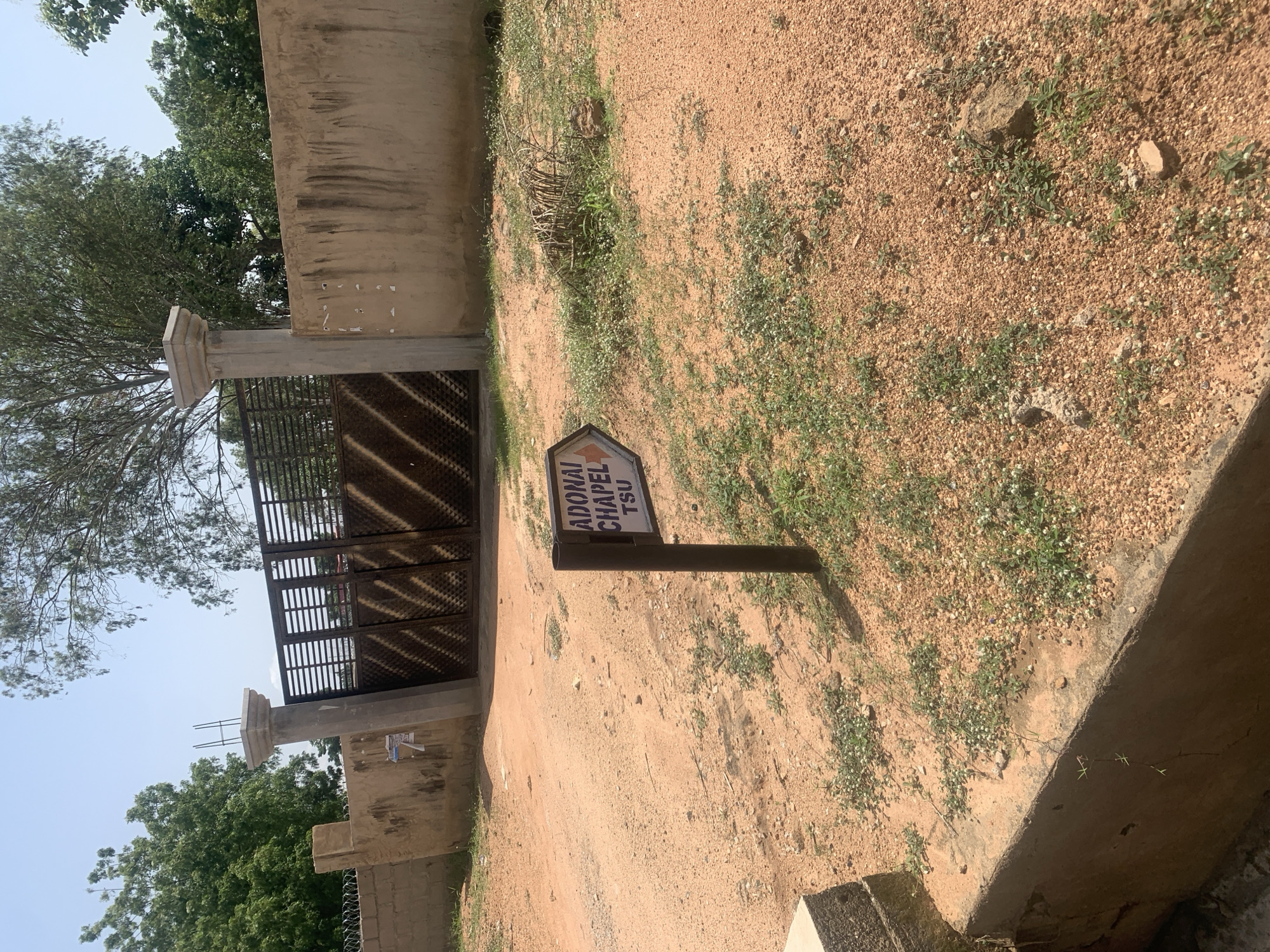 The image depicts an outdoor scene in Jalingo, Nigeria, featuring a dirt road leading to a large metal gate and a concrete wall. A signpost with an arrow pointing to the right, indicating "ADONAI CHAPEL TSU," is prominently placed in the foreground. The ground is covered with reddish-brown dirt, small pebbles, and sparse green vegetation. Behind the signpost, a concrete structure or planter box is visible on the left. The gate, made of dark metal with a diamond pattern, is closed and appears to be the entrance to a compound. The wall is a light brown color, with some marks and peeling paint. Trees with green foliage and a clear blue sky are visible in the background. The overall impression is of a slightly arid, sun-drenched environment.