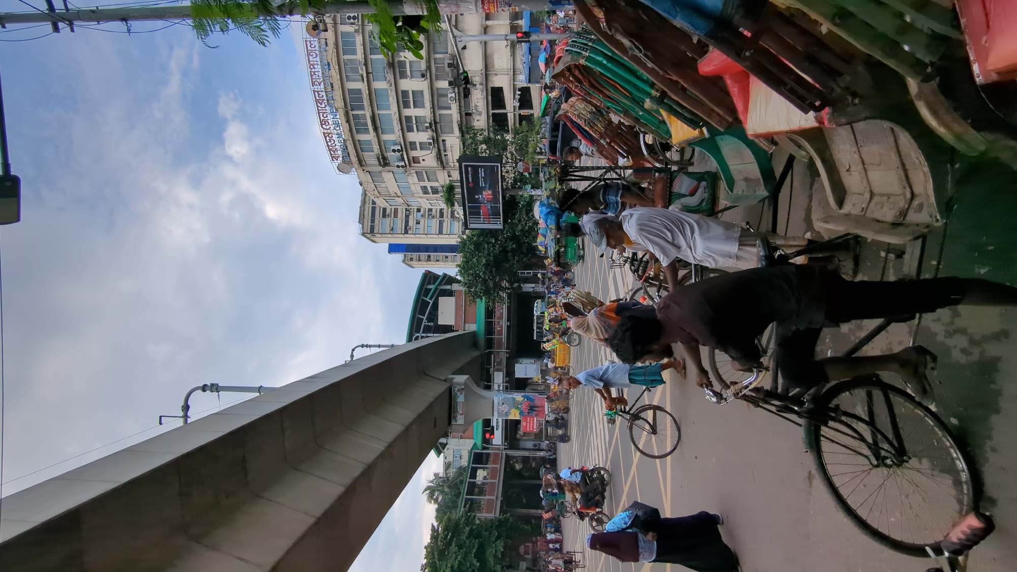 The image depicts a busy street intersection in Dhaka, Bangladesh, under an elevated metro line. Several people are present, including pedestrians, cyclists, and rickshaw pullers. In the foreground, a man in a dark shirt is operating a bicycle rickshaw. To his right, several other rickshaws are parked, some with passengers. In the background, a building with signage in Bengali and English reads "Kurmitola General Hospital." Traffic, including motorcycles and other vehicles, can be seen moving through the intersection. The sky is partly cloudy, and the overall scene suggests a typical day in a bustling urban environment.